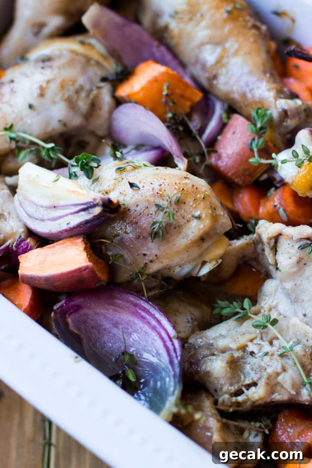 White dish containing oven baked chicken with sweet potatoes, carrots, and red onions topped with fresh thyme.