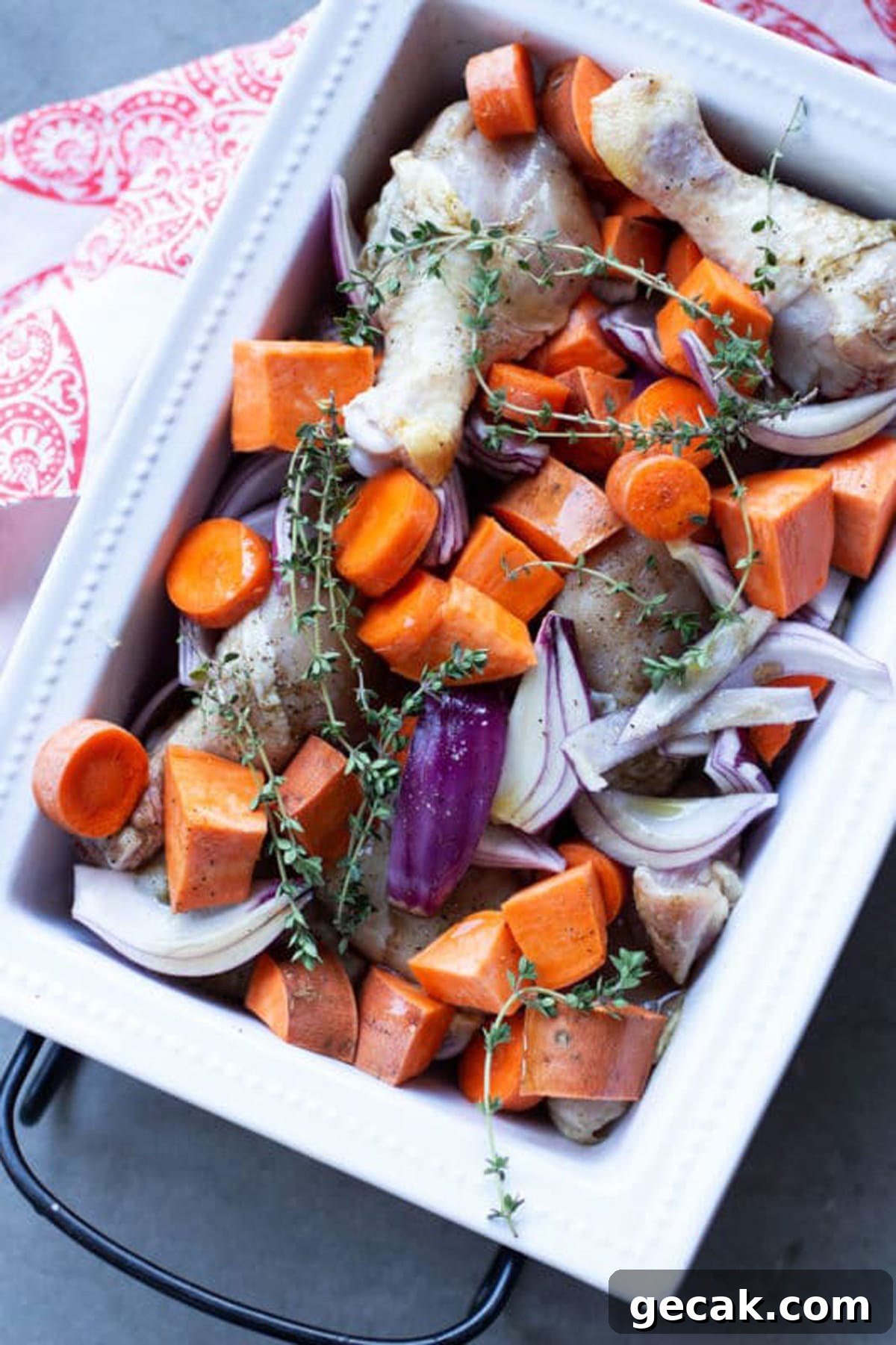 White dish containing seasoned drumsticks with sweet potatoes, carrots, and red onions topped with fresh thyme.