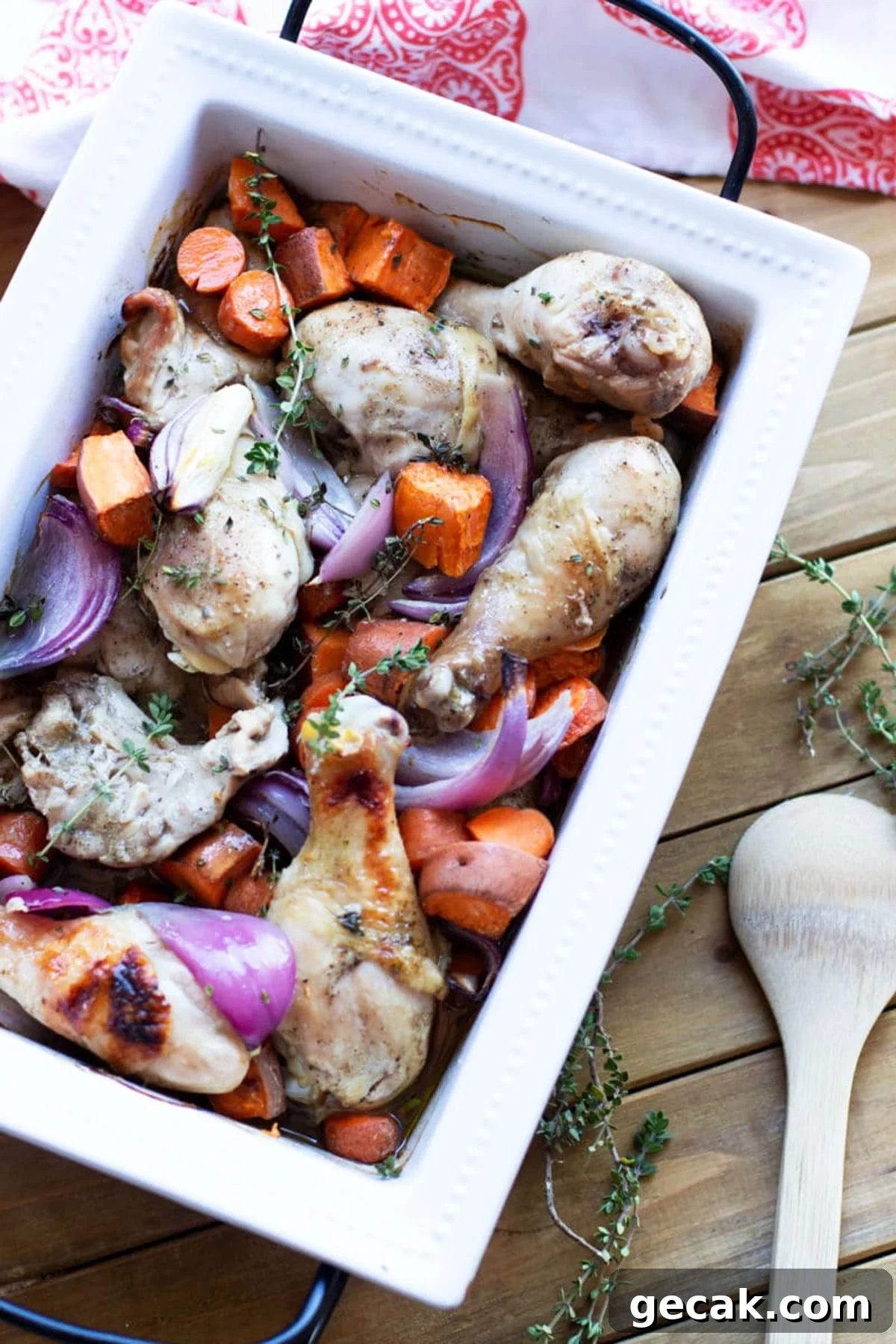 White dish containing roasted chicken drumsticks with sweet potatoes, carrots, and red onions topped with fresh thyme.