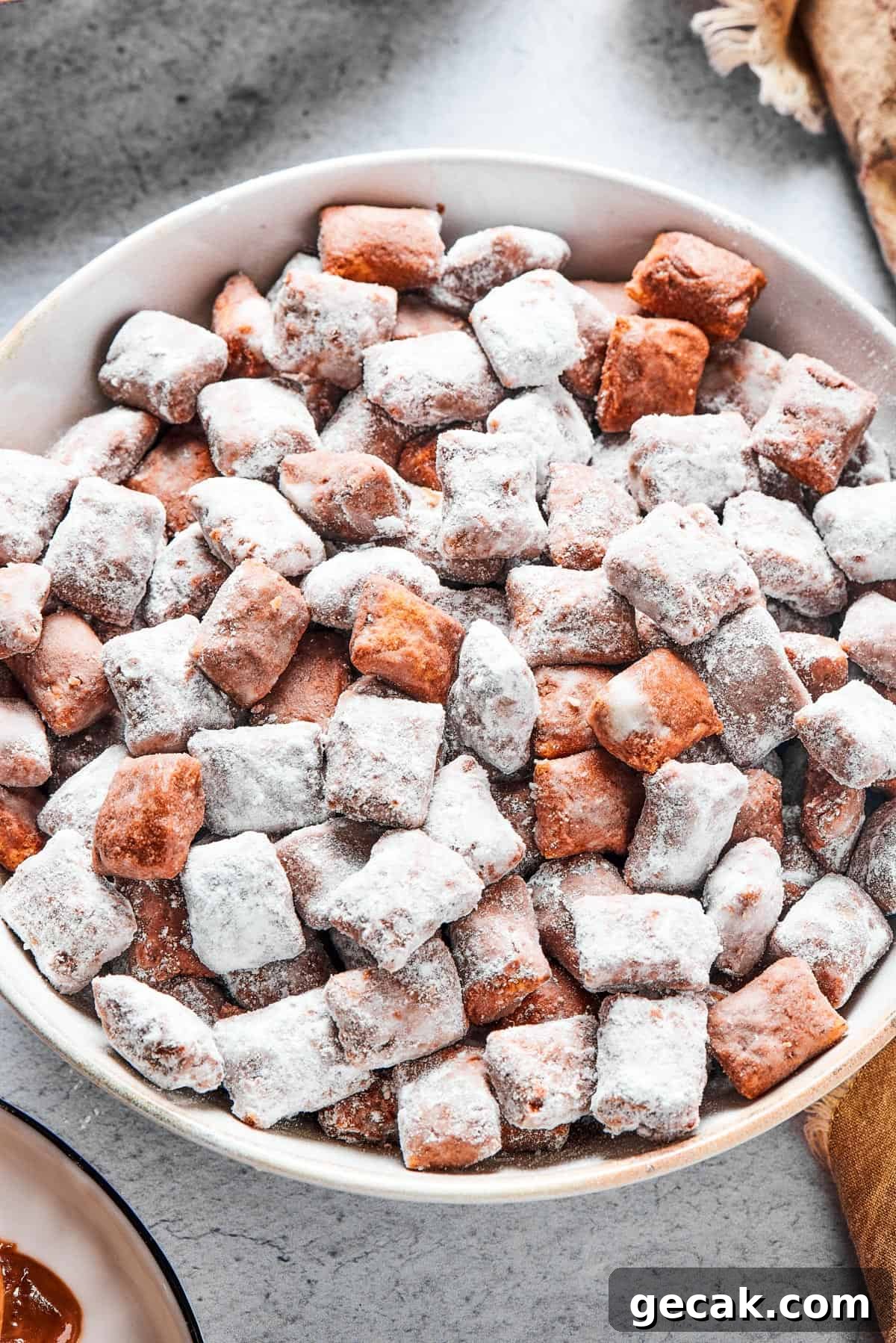 Who’s up for Muddy Buddies? Smooth chocolate, salty peanut butter, and a light dusting of powdered sugar make this crispy Chex cereal snack a true favorite! Close-up shot of puppy chow in a white bowl, to show the texture. Some pieces are fully coated in powdered sugar, while others are not.