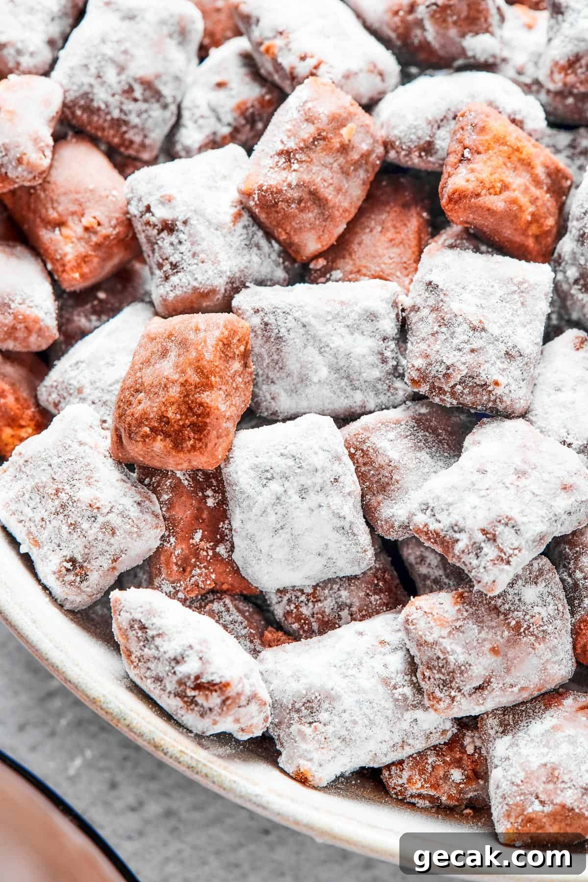 Who’s up for Muddy Buddies? Smooth chocolate, salty peanut butter, and a light dusting of powdered sugar make this crispy Chex cereal snack a true favorite! Close-up shot of puppy chow in a white bowl, to show the texture. Some pieces are fully coated in powdered sugar, while others are not.
