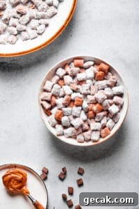 Chocolate Peanut Butter Muddy Buddies Snack Mix Recipe 7 Muddy buddies in a white bowl, with a spoonful of peanut butter and a larger serving dish of the snack nearby.