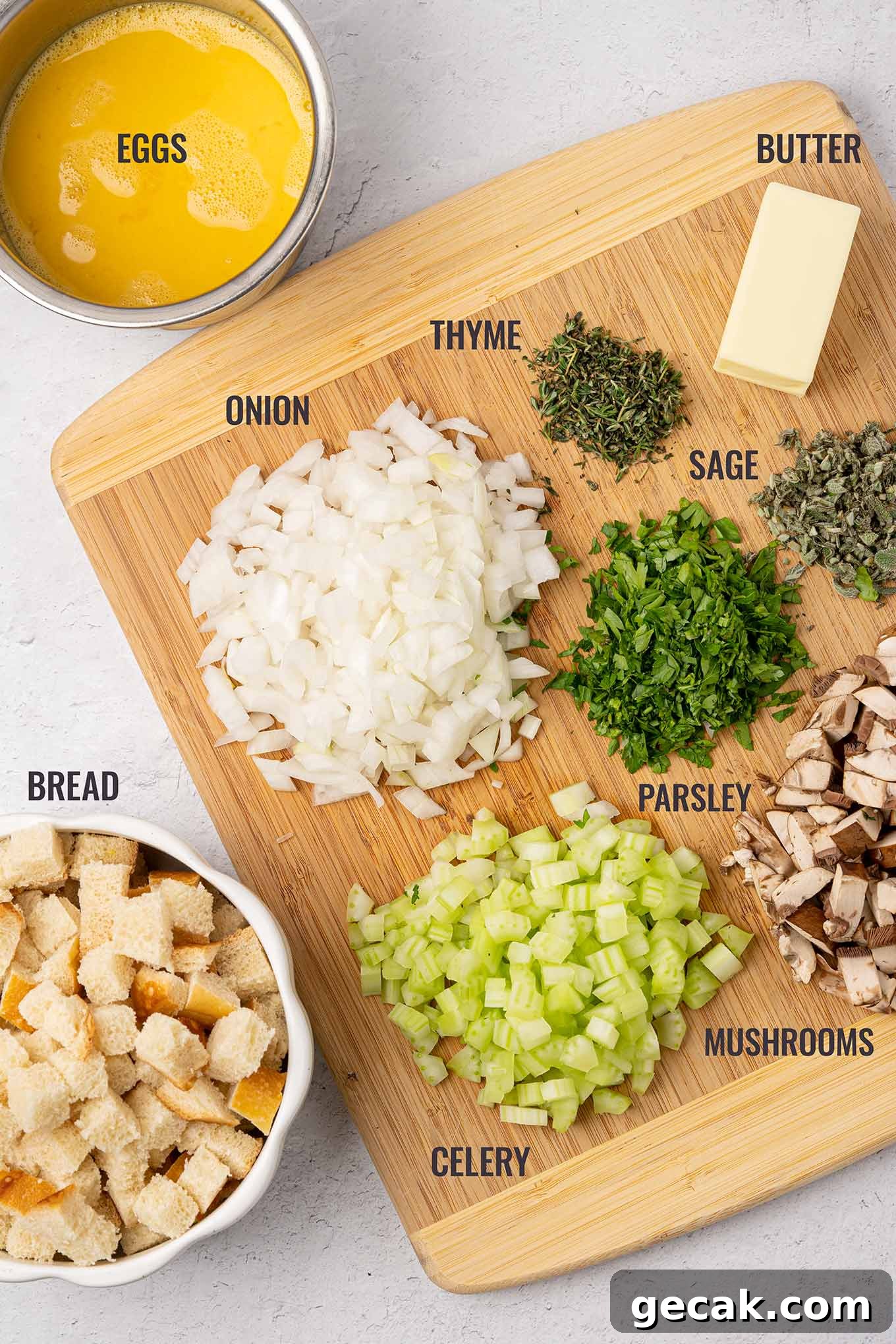 Classic Old-Fashioned Bread Stuffing Recipe for Thanksgiving 3 A labeled, overhead view of the ingredients for bread stuffing including bread cubes, eggs, butter, onion, celery, mushrooms, fresh thyme, parsley, and sage.