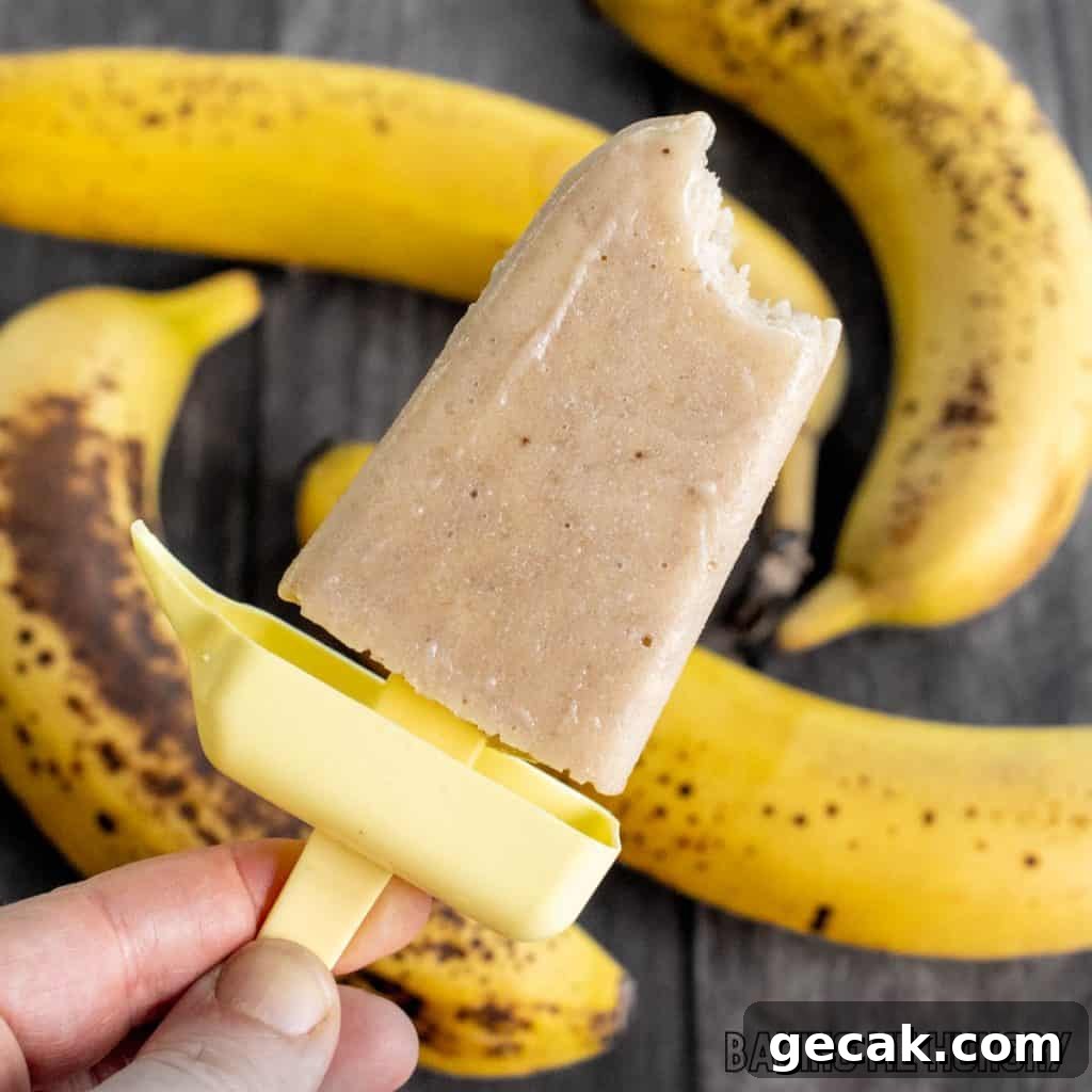 creamy banana popsicles recipe shown as a hand holding up a pop missing a bite with bananas behind