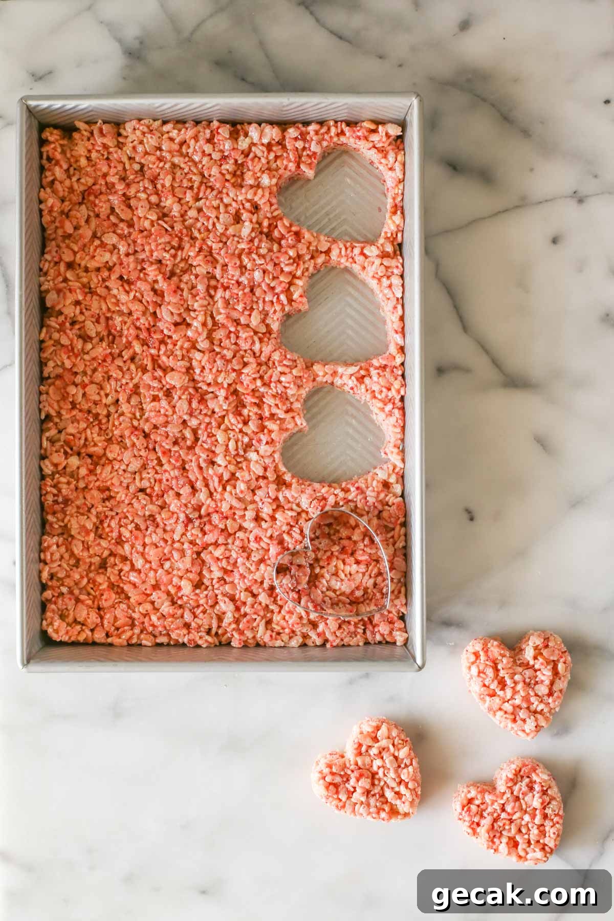 Strawberry Rice Krispie Treats Recipe – Fresh Fruity Snack 2 Overhead shot of a pan of Strawberry Rice Krispie Treats with a heart shaped cutter in the pan with three hearts already cut out and laying next to the pan, all on a marble background.