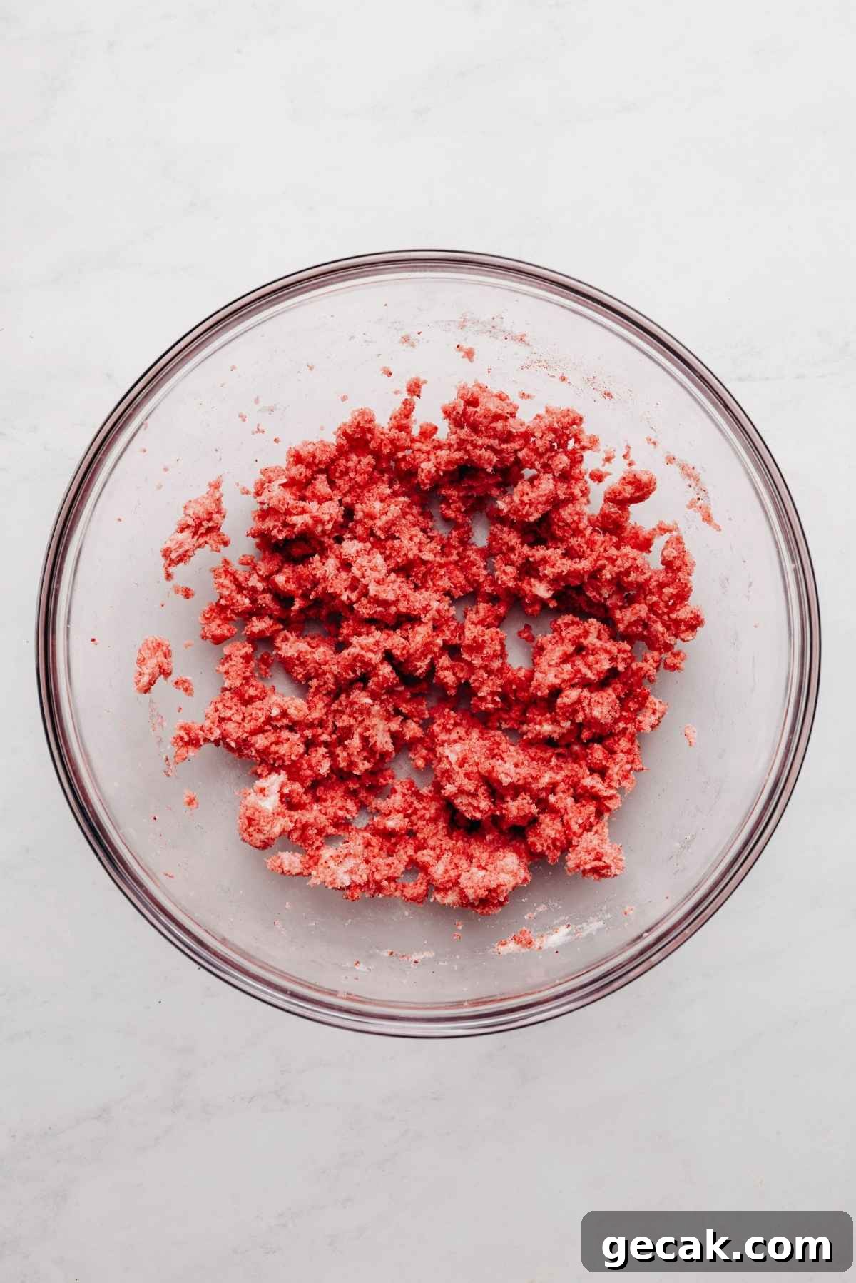 Freeze dried strawberry powder, butter, and sugar blended together in a glass mixing bowl on a white and grey textured background.