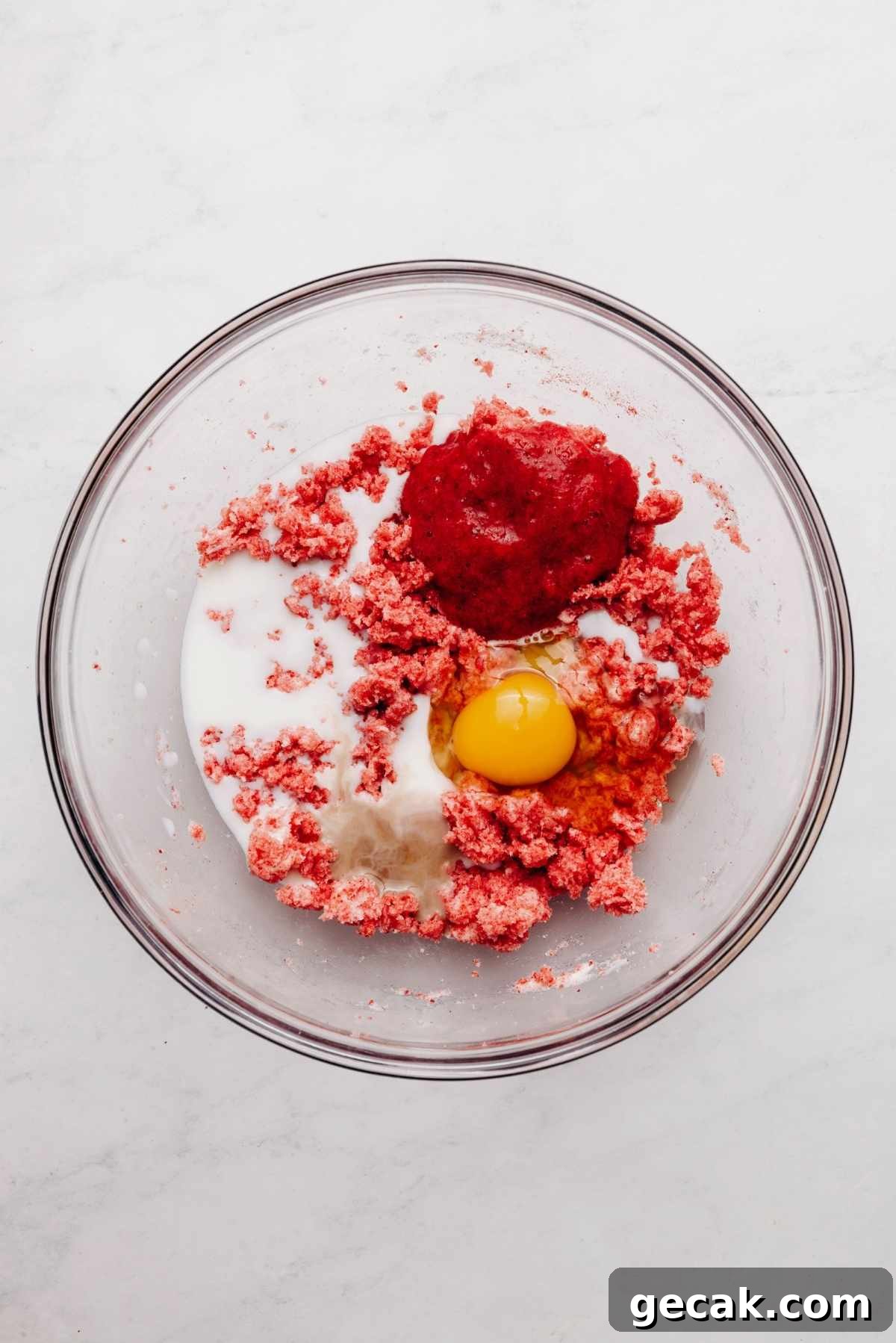 Vanilla extract, milk, roasted strawberry puree, and an egg added to the top of the cake mixture in a glass mixing bowl.