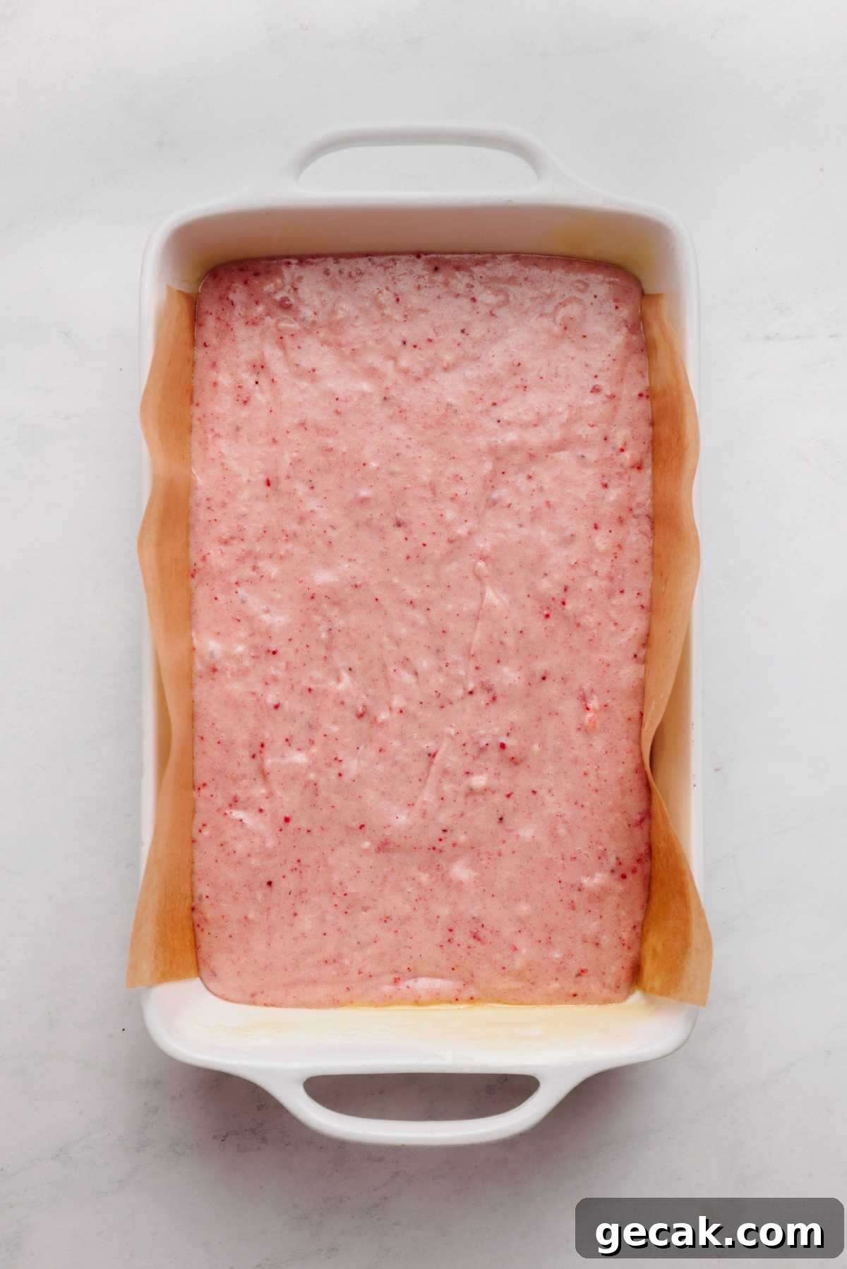 Strawberry cake batter in a parchment paper lined ceramic baking tin on a white and grey textured background.
