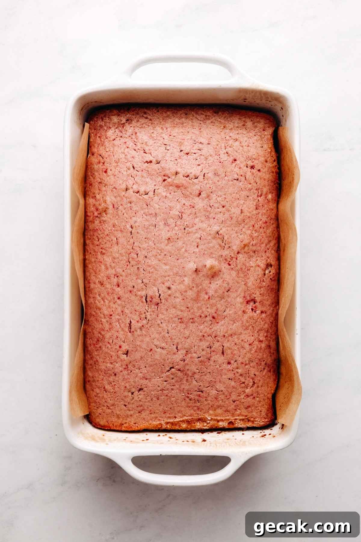 Baked mini strawberry sheet cake in a parchment paper lined baking tin on a white and grey textured background.