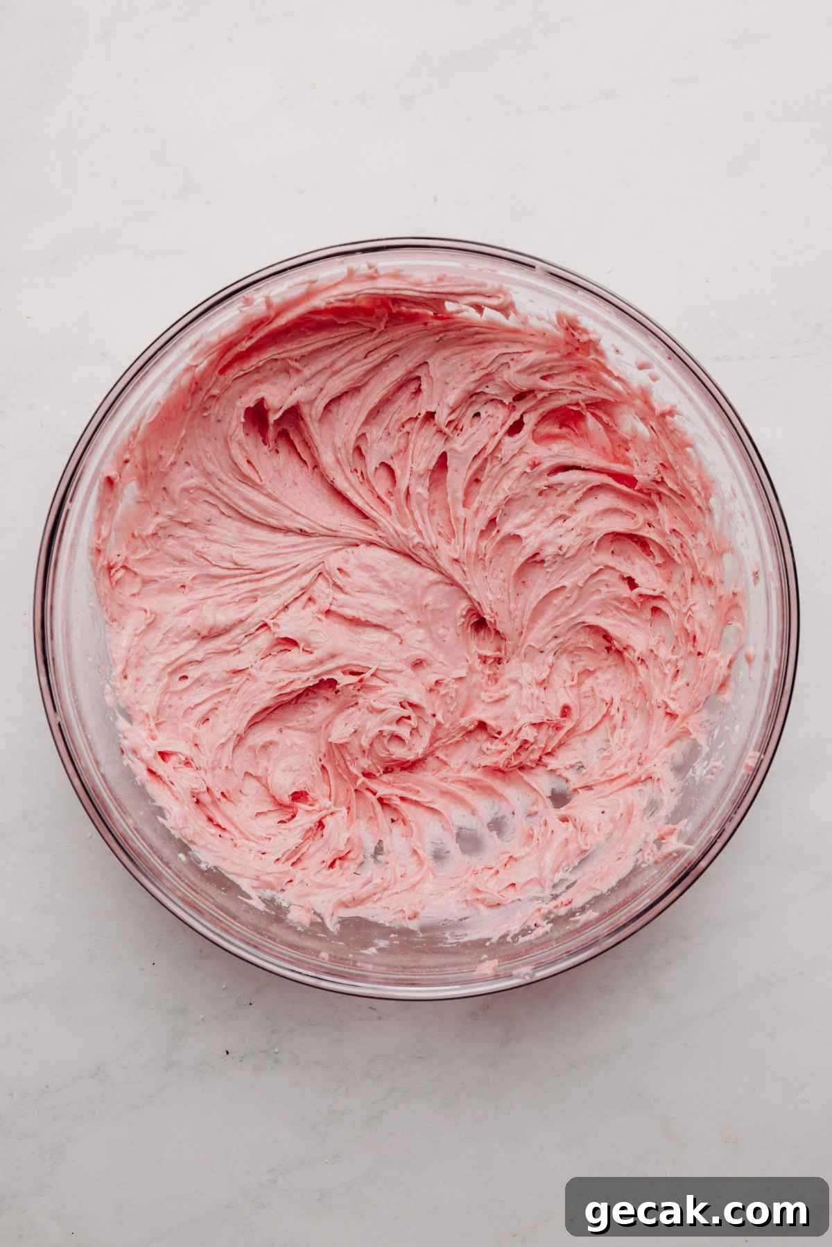 Strawberry buttercream frosting in a large glass mixing bowl on a white and grey textured background.