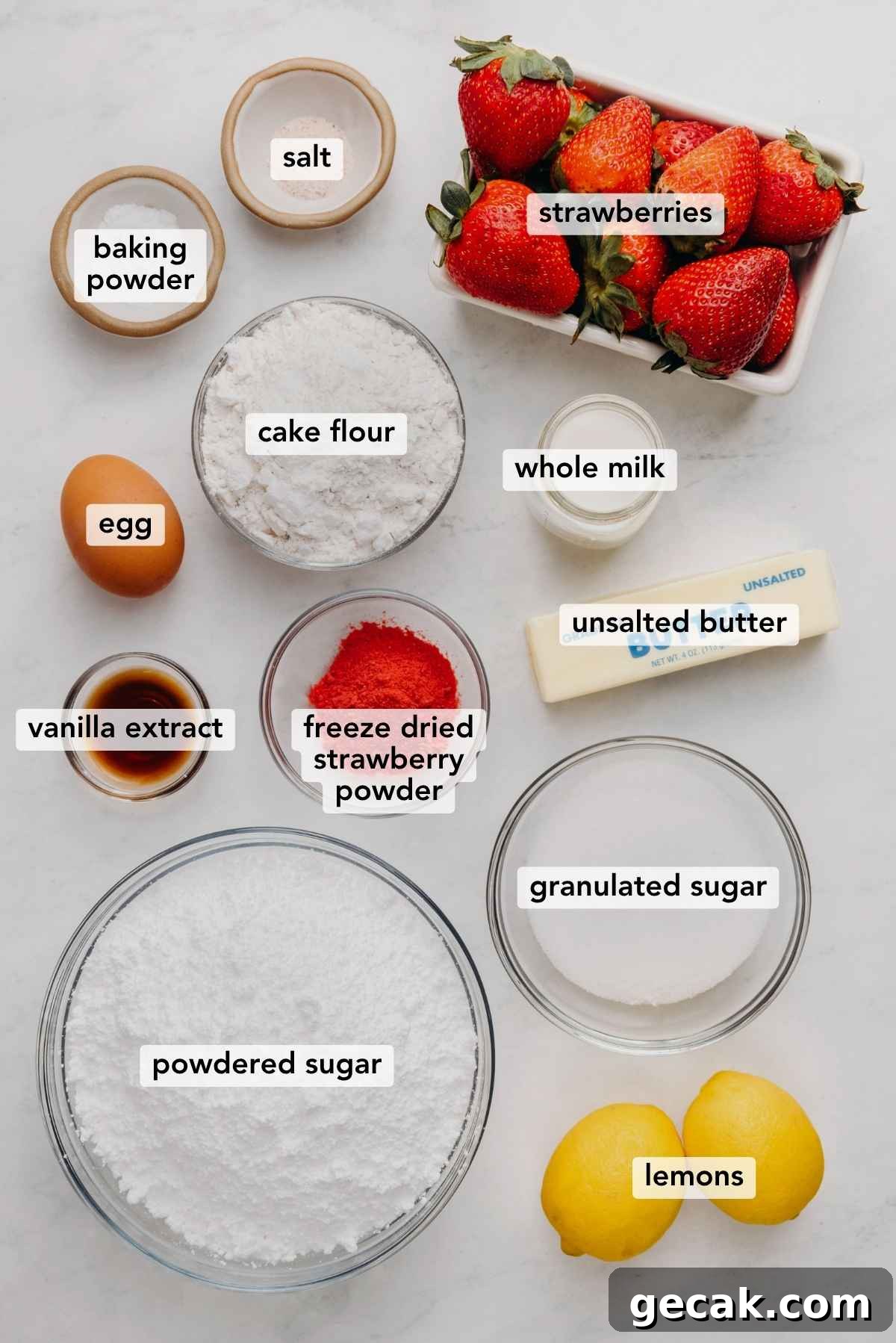 Ingredients in various size bowls labeled as baking powder, salt, strawberries, egg, cake flour, whole milk, vanilla extract, freeze dried strawberry powder, unsalted butter, powdered sugar, granulated sugar, and lemons on a white marbled background.