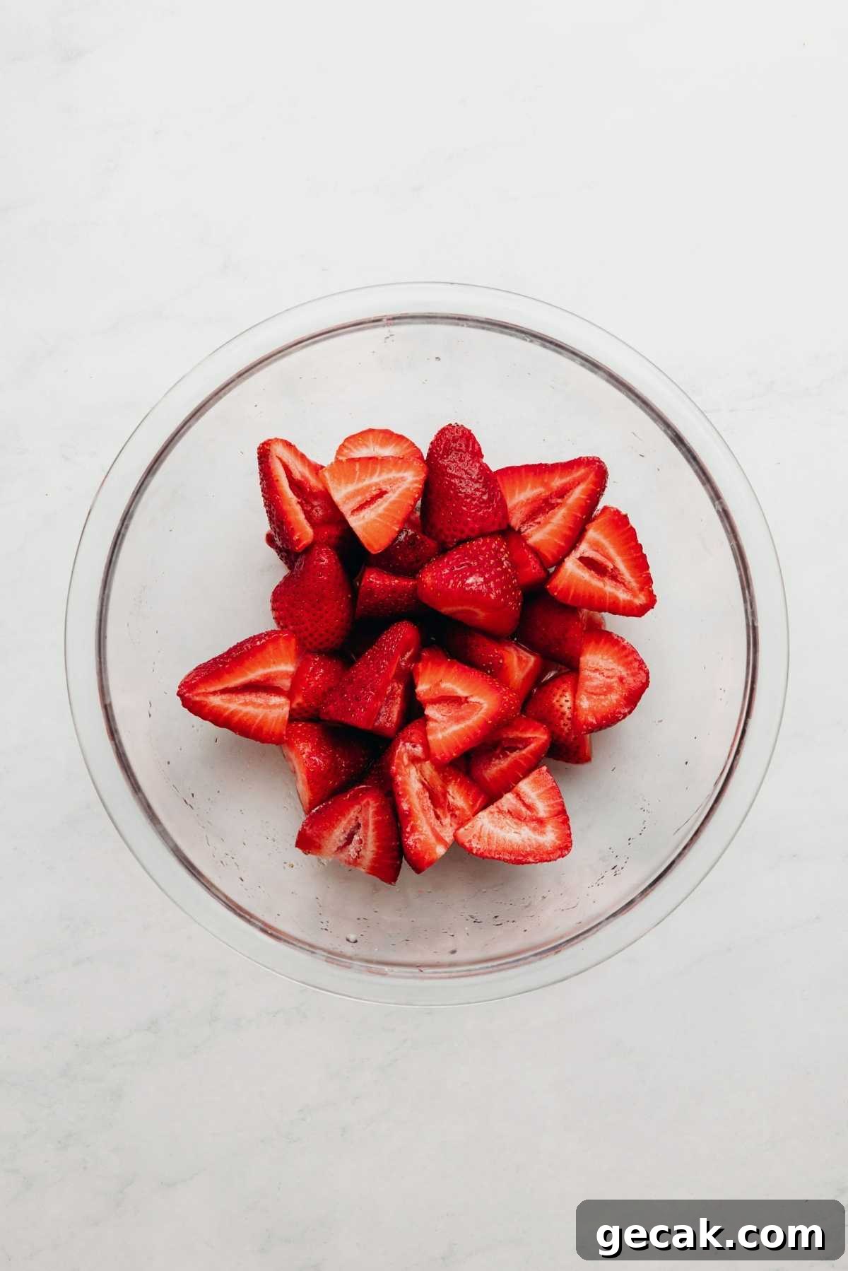 Sliced strawberries, lemon juice, and granulated sugar tossed together in a small glass bowl on a white and grey textured background.