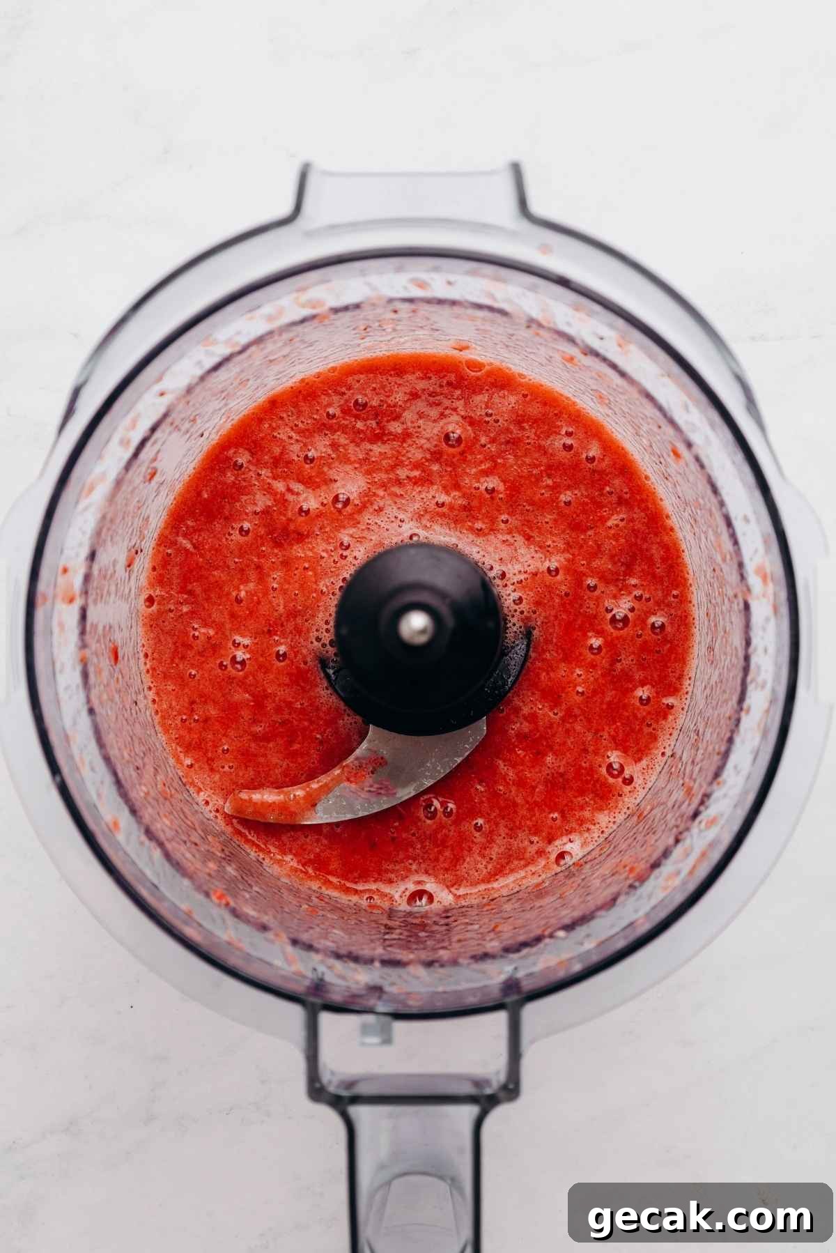Roasted strawberry puree in a food processor on a white and grey textured background.