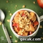 Overhead shot of apple pie snack in a white mug with a spoon and graham cracker crumbles on the side all on a green surface.