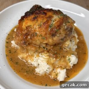 A plate of mashed potatoes topped with a browned, saucy Smothered Chicken thigh and garnished with herbs, with extra orange-brown gravy pooling around the base. A pot is visible in the background on a wooden surface.