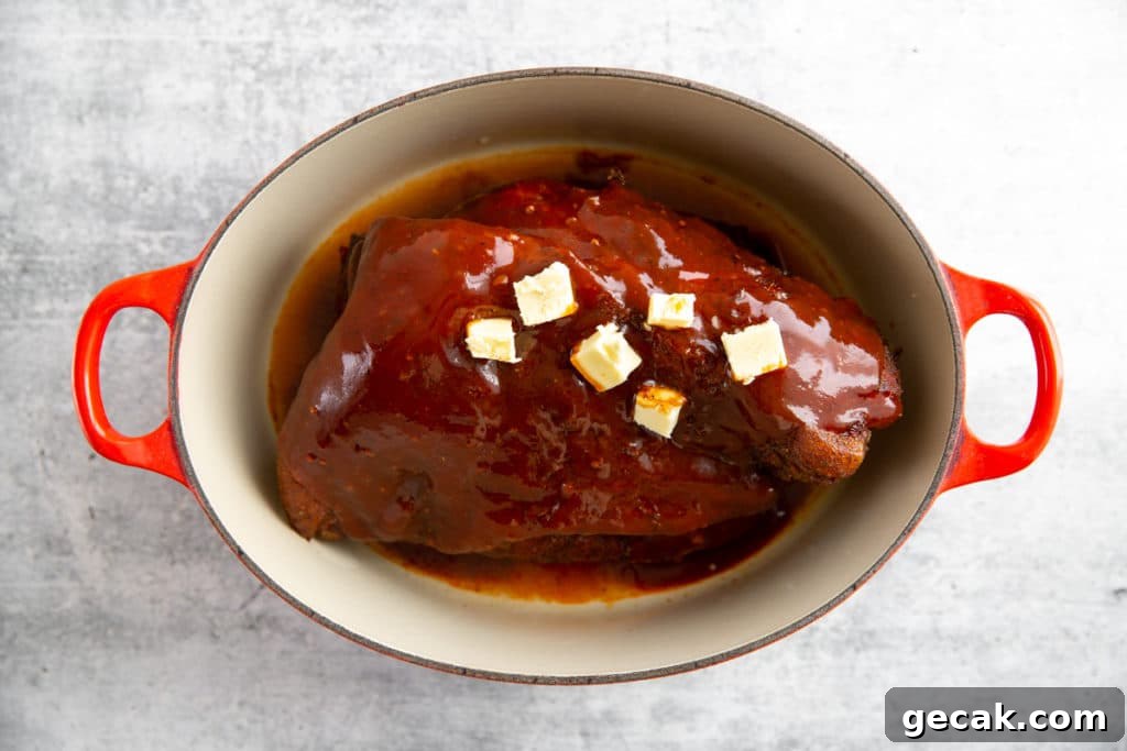 Process shot showing a smoked pork butt in a dutch oven, topped with barbecue sauce and butter.