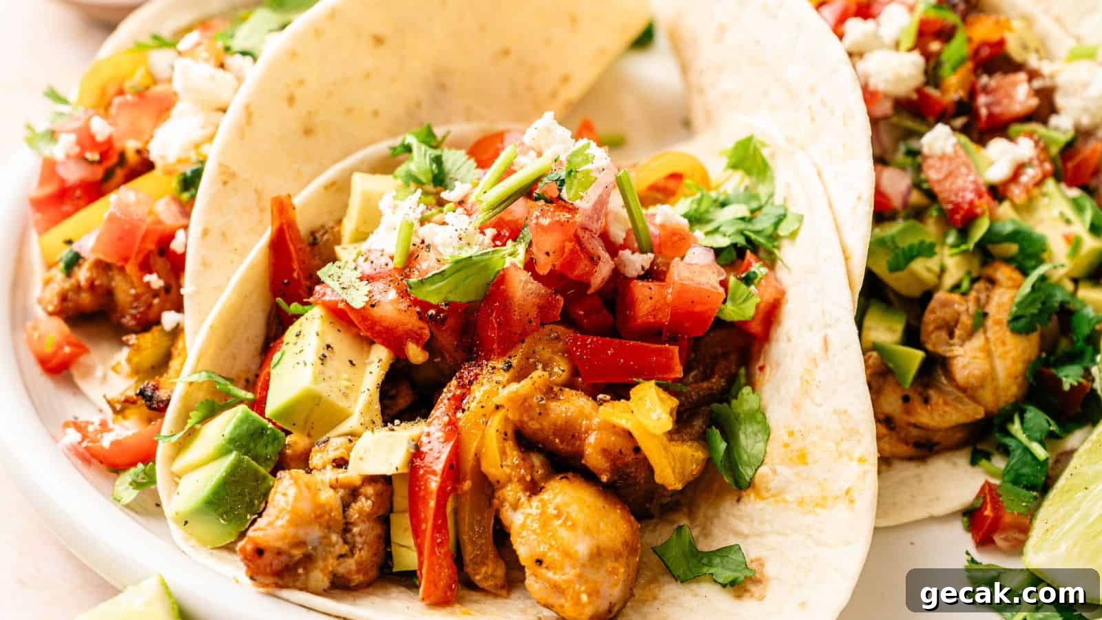 Three soft tacos filled with seasoned chicken, diced avocado, chopped tomatoes, crumbled cheese, and fresh cilantro on a white plate.