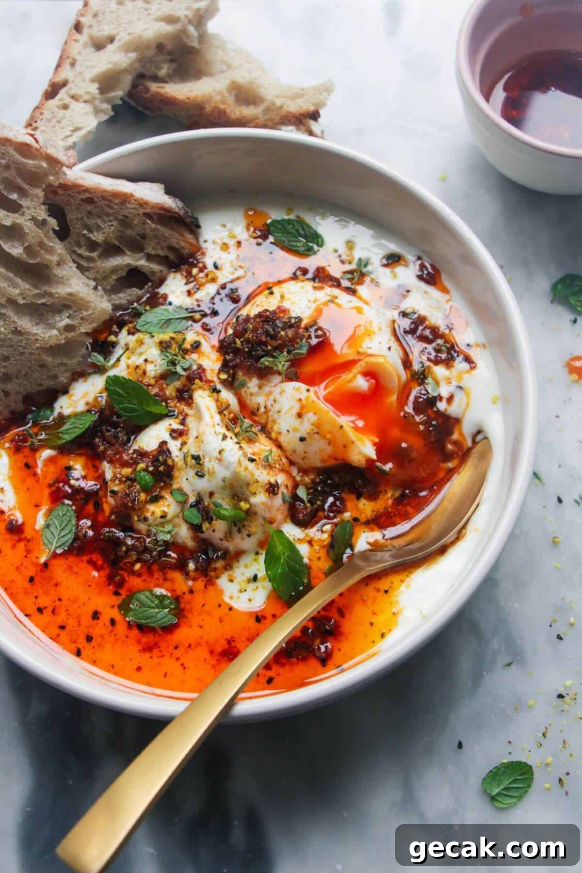 Turkish-Style Poached Eggs with Tomato and Yogurt Sauce 2 Small white bowl with 2 poached eggs, 1 broken open with yolk coming out, with a gold spoon on the side and toast in the bowl.