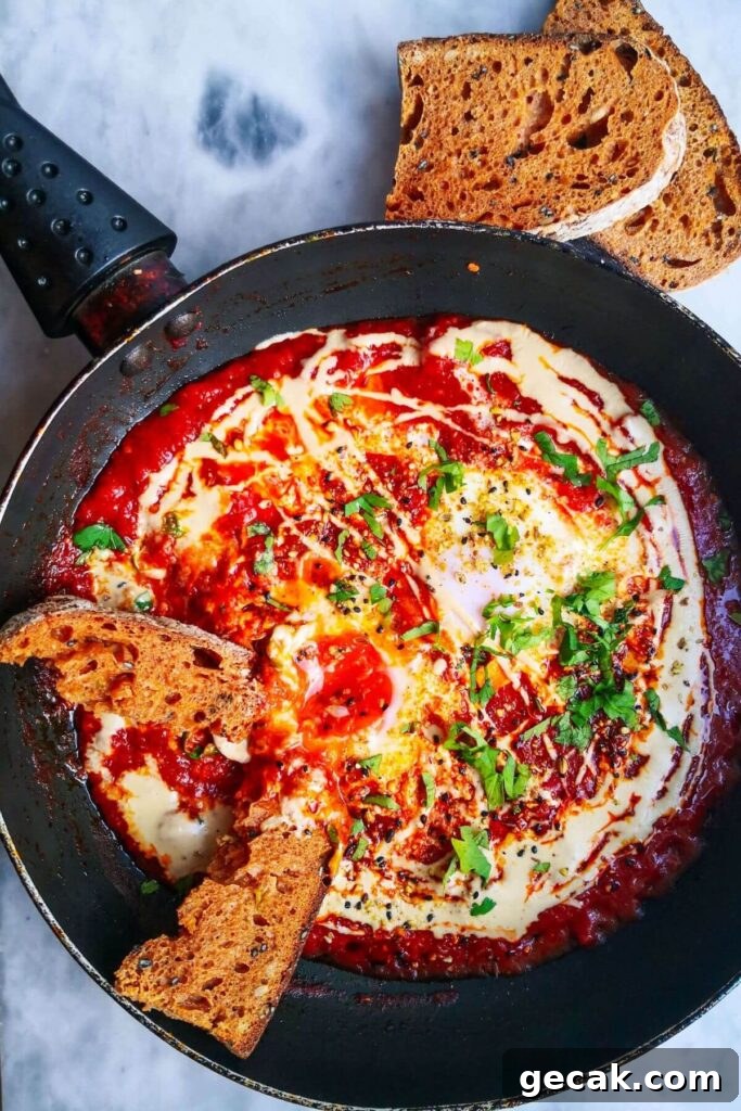 Turkish-Style Poached Eggs with Tomato and Yogurt Sauce 14 Shakshuka for one in a small black pan, on a grey marble background with sourdough placed into the sauce, and two slices of sourdough on the side.