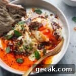 Small white bowl with 2 poached eggs, 1 broken open with yolk coming out, with a gold spoon on the side and toast in the bowl.