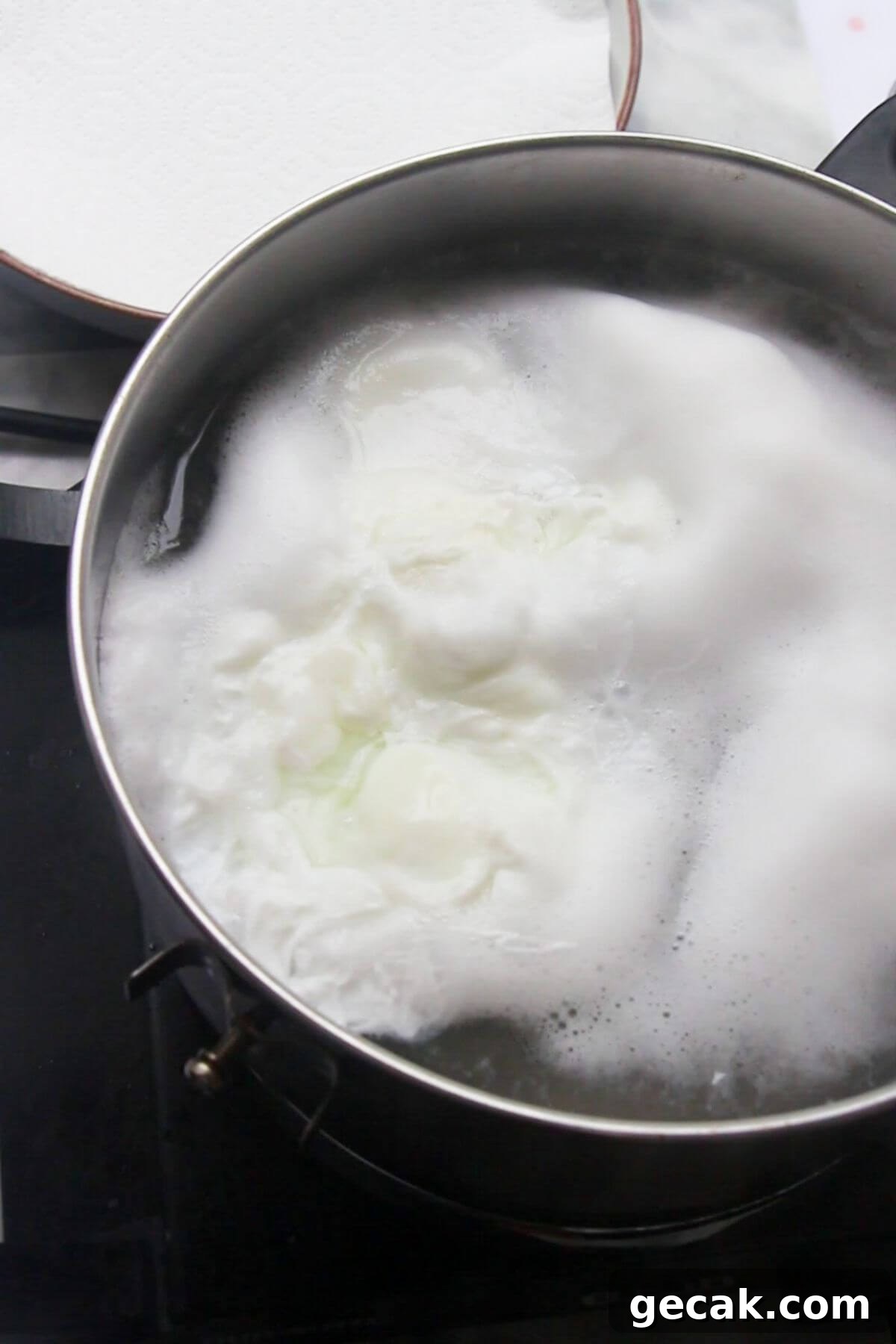 Turkish-Style Poached Eggs with Tomato and Yogurt Sauce 8 2 eggs poaching in a small silver pot with lined plate in the background.