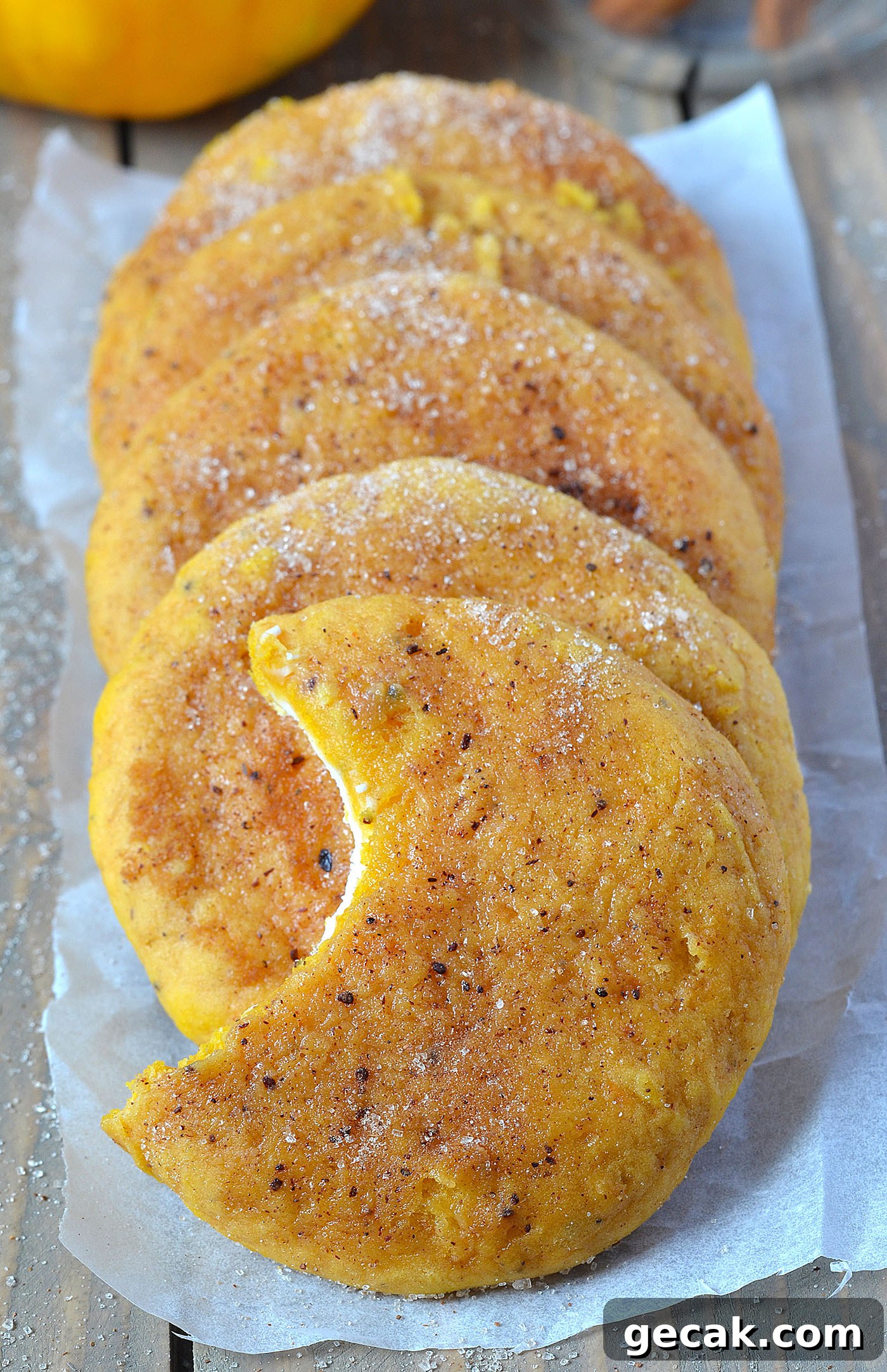 Row of Snickerdoodle Cream Cheese Pumpkin Cookies arranged on parchment paper, each cookie golden brown with cinnamon sugar coating and one cookie showing a bite revealing the creamy filling inside.