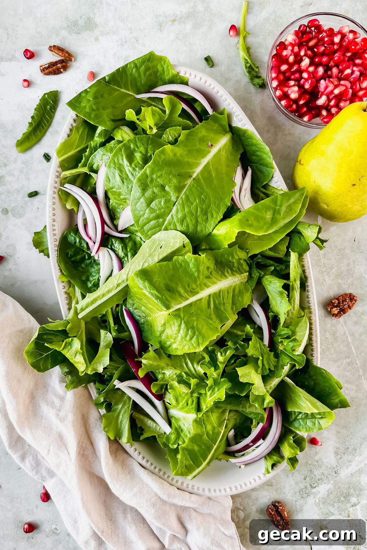 lettuce and red onion on white serving plate beside small bowl of pomegranate seeds and a pear.