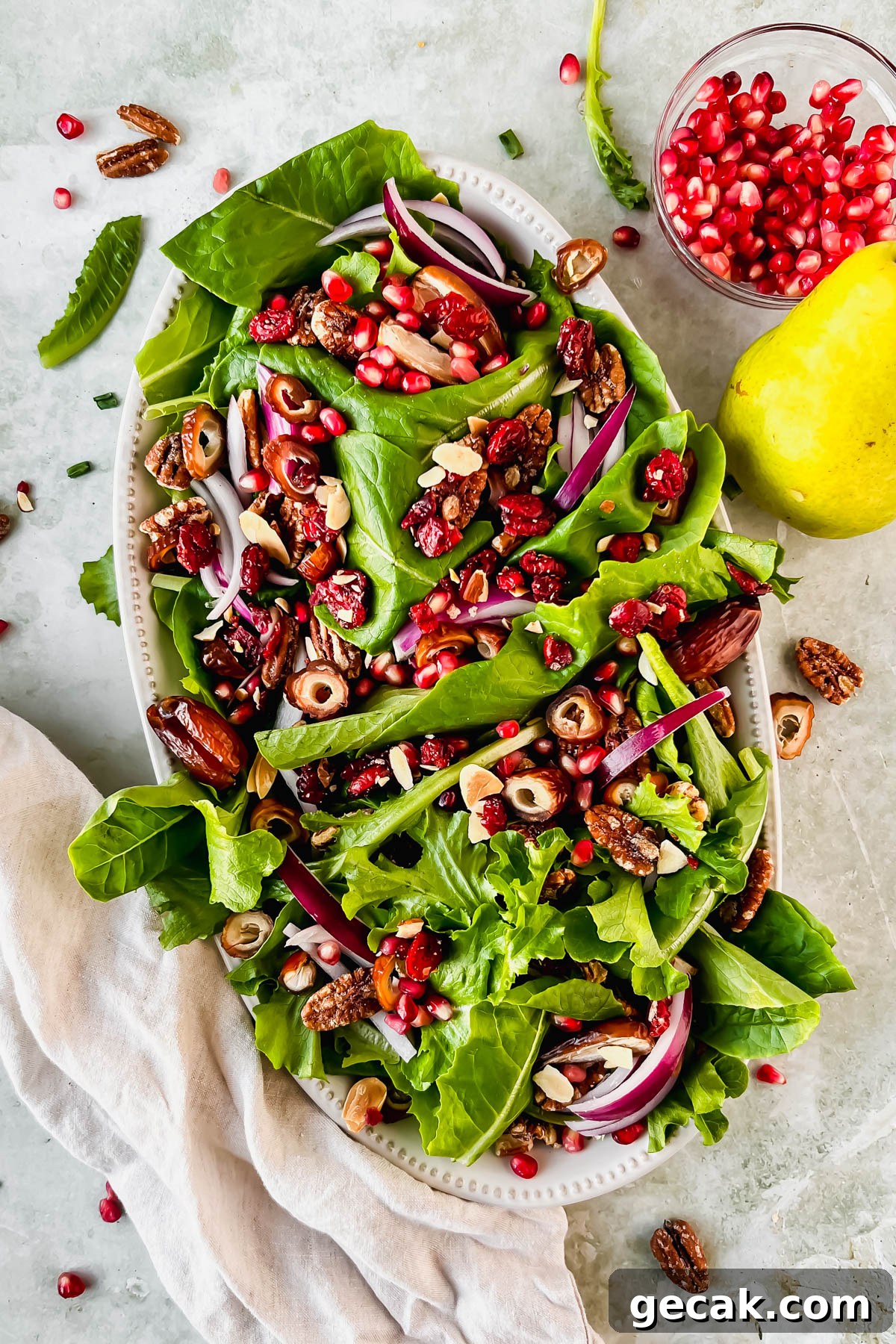 lettuce, sliced dates, pecans and red onion on white serving plate beside small bowl of pomegranate seeds and a pear.
