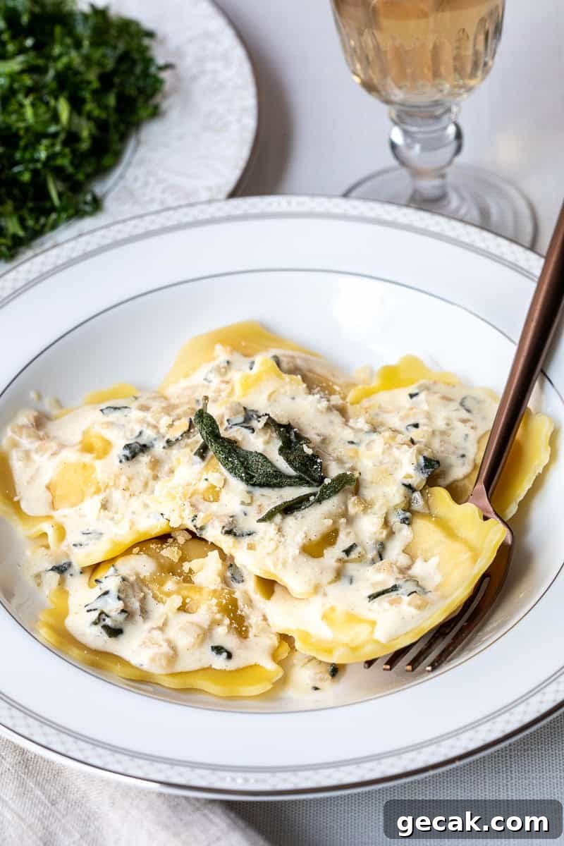 A white plate of lobster ravioli topped with creamy sauce, grated cheese, and sage leaves, with a fork and a glass of white wine nearby.