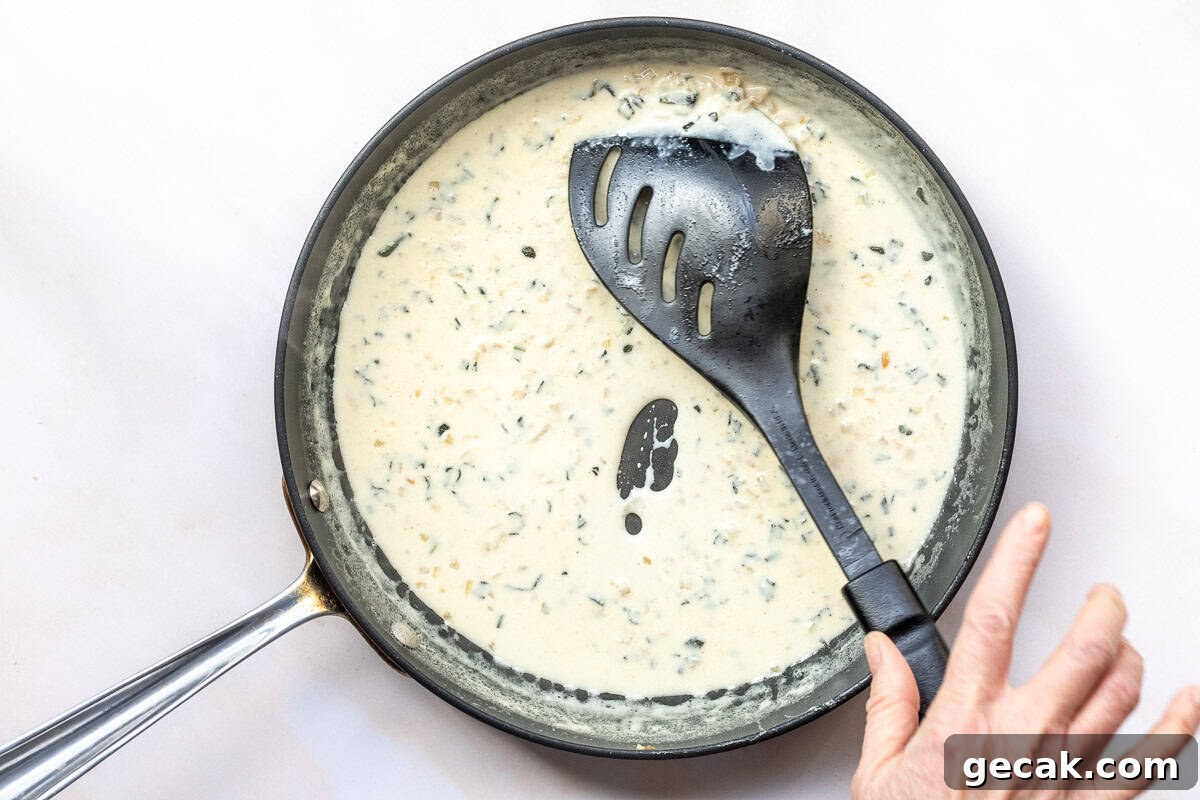 A black frying pan with creamy lobster ravioli sauce and herbs, stirred with a black slotted spatula. A hand is visible near the edge of the pan.