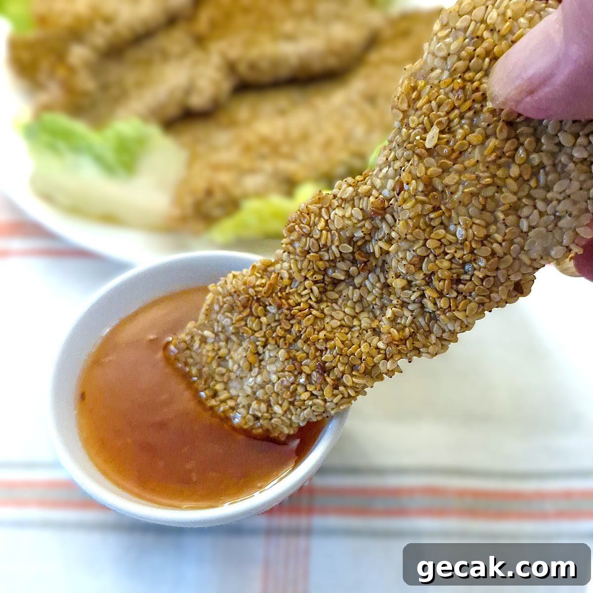 Crispy Sesame Chicken Strips Without Breadcrumbs — Low-Carb Crunch 2 A sesame-coated chicken goujon being dipped into a bowl of dipping sauce.