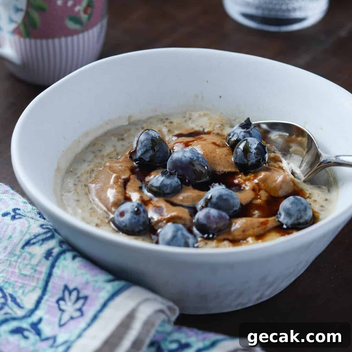photo of a white bowl of vegan oat and chia seed porridge with blueberries and coconut almond butter on top, all on a dark wood table with a napkin to the side and cup of coffee in the background.