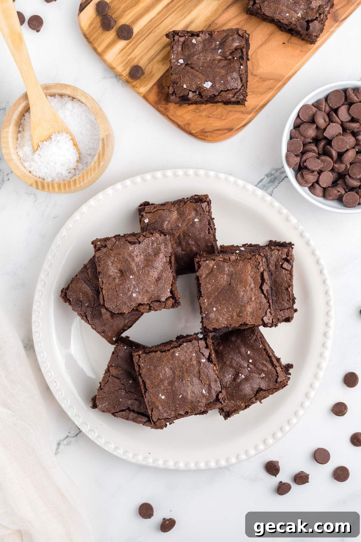 A plate of homemade brownies on a white surface with chocolate chips and a stack of brownies nearby; a fork and napkin are beside the plate.