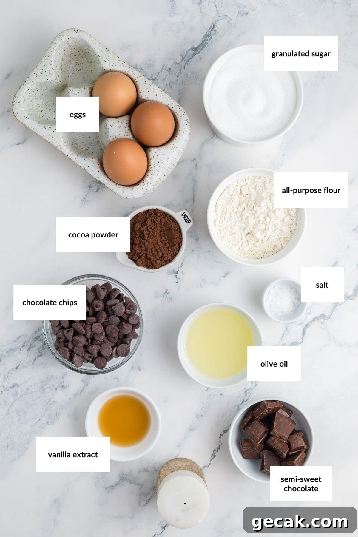 Ingredients for baking homemade brownies are arranged on a marble surface, including eggs, sugar, flour, cocoa powder, salt, olive oil, chocolate chips, vanilla extract, and semi-sweet chocolate.