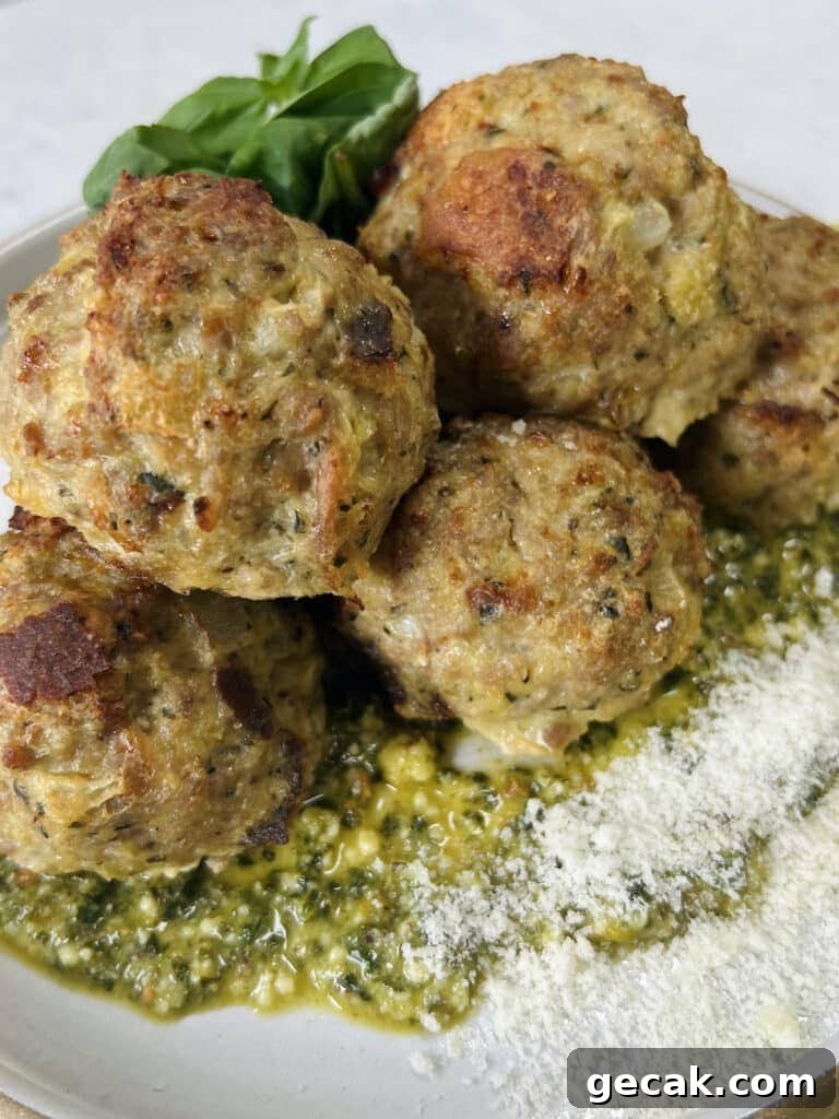 Styled close up of cooked turkey meatballs with pesto on top of pesto on plate, garnished with grated cheese and whole basil leaves.