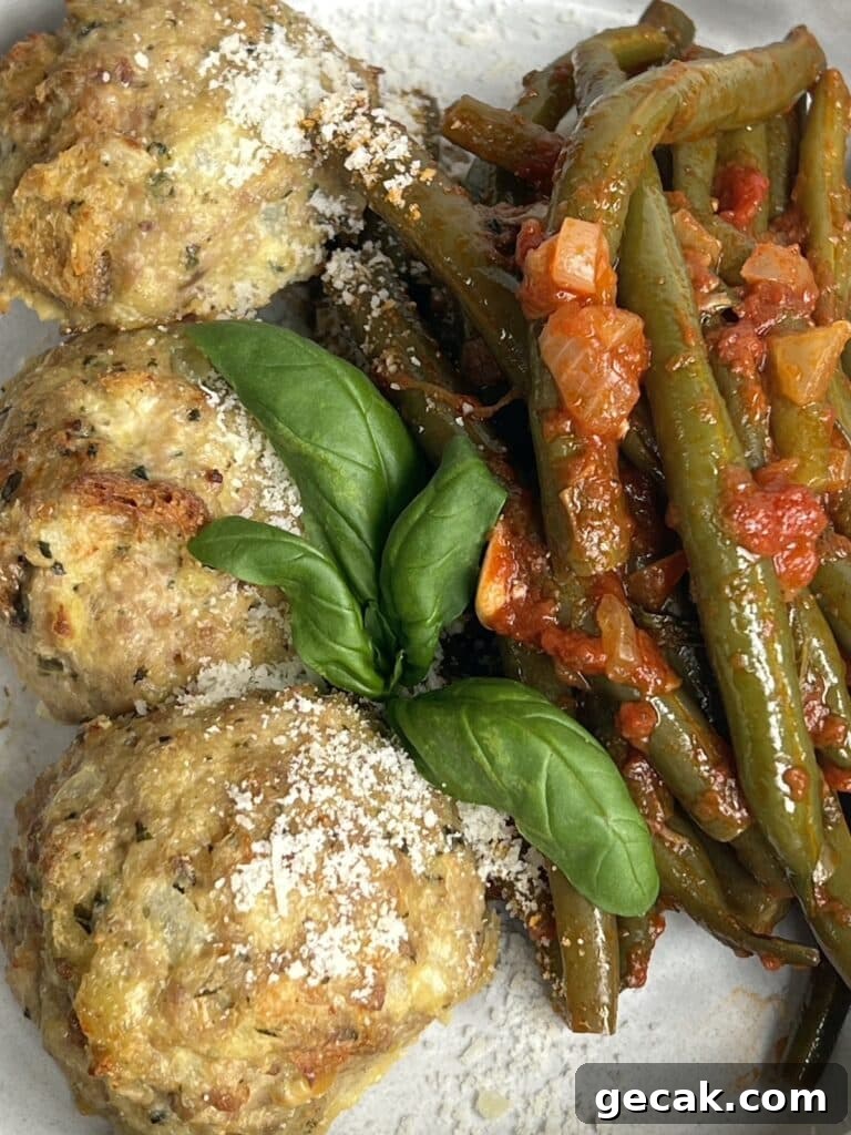Overhead view of three cooked turkey meatballs with pesto on plate with green beans in tomato sauce and basil garnish.