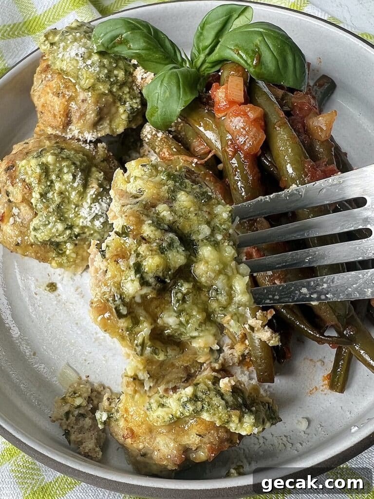 Close up of cooked turkey meatballs with pesto on fork with green beans in tomato sauce and basil garnish, topped with grated cheese.