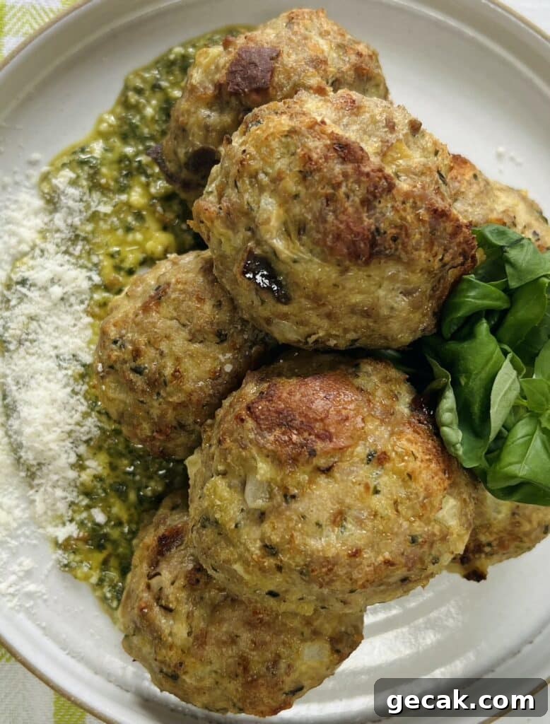 Overhead view of styled close up of cooked turkey meatballs with pesto on top of pesto on plate, garnished with grated cheese and whole basil leaves.