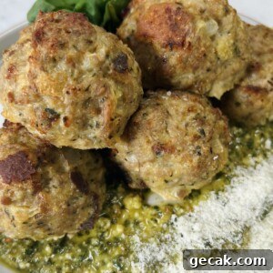 Styled close up of cooked turkey meatballs with pesto on top of pesto on plate, garnished with grated cheese and whole basil leaves.