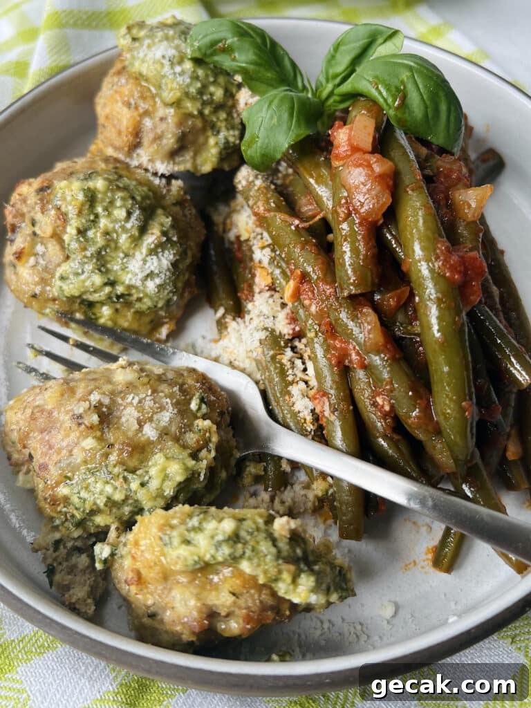 Overhead view of three cooked turkey meatballs with pesto on plate with green beans in tomato sauce and basil garnish, topped with grated cheese.