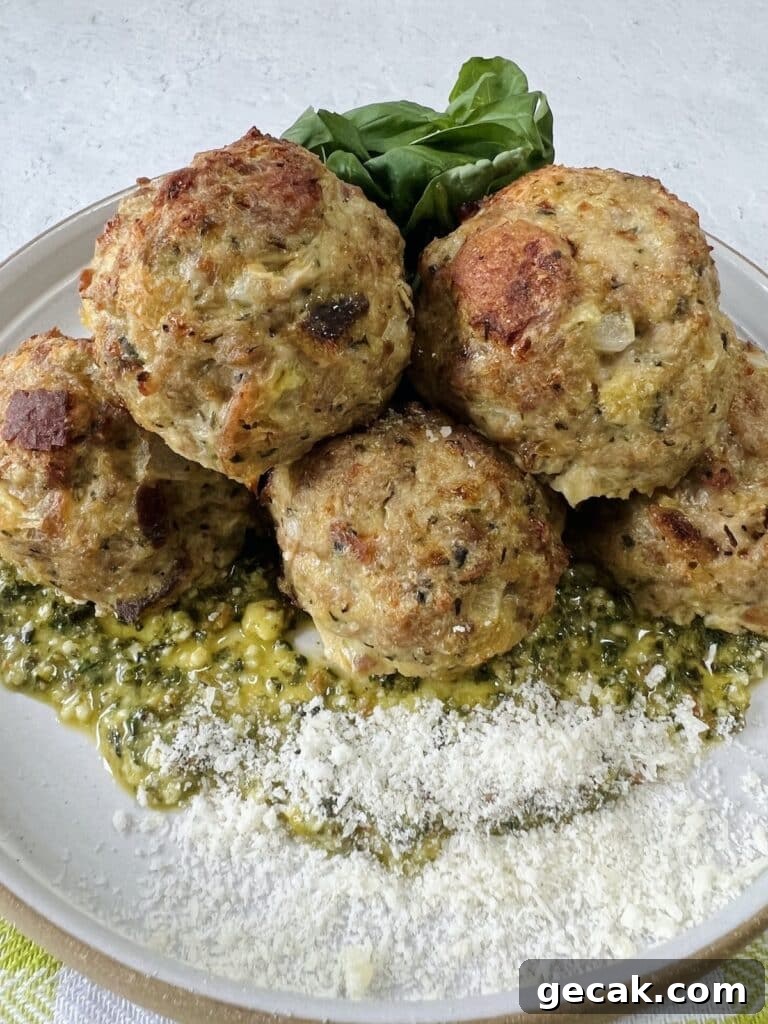 Styled close up of cooked turkey meatballs with pesto on top of pesto on plate, garnished with grated cheese and whole basil leaves.