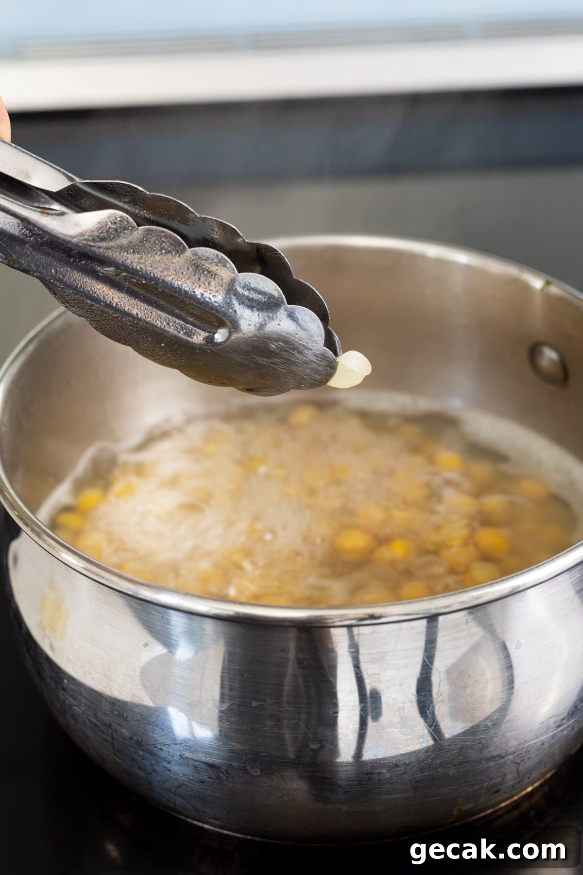 Creamy Chickpea Hummus Recipe: Classic Homemade Dip 3 Tongs holding a chickpea skin above a pot of cooking chickpeas.