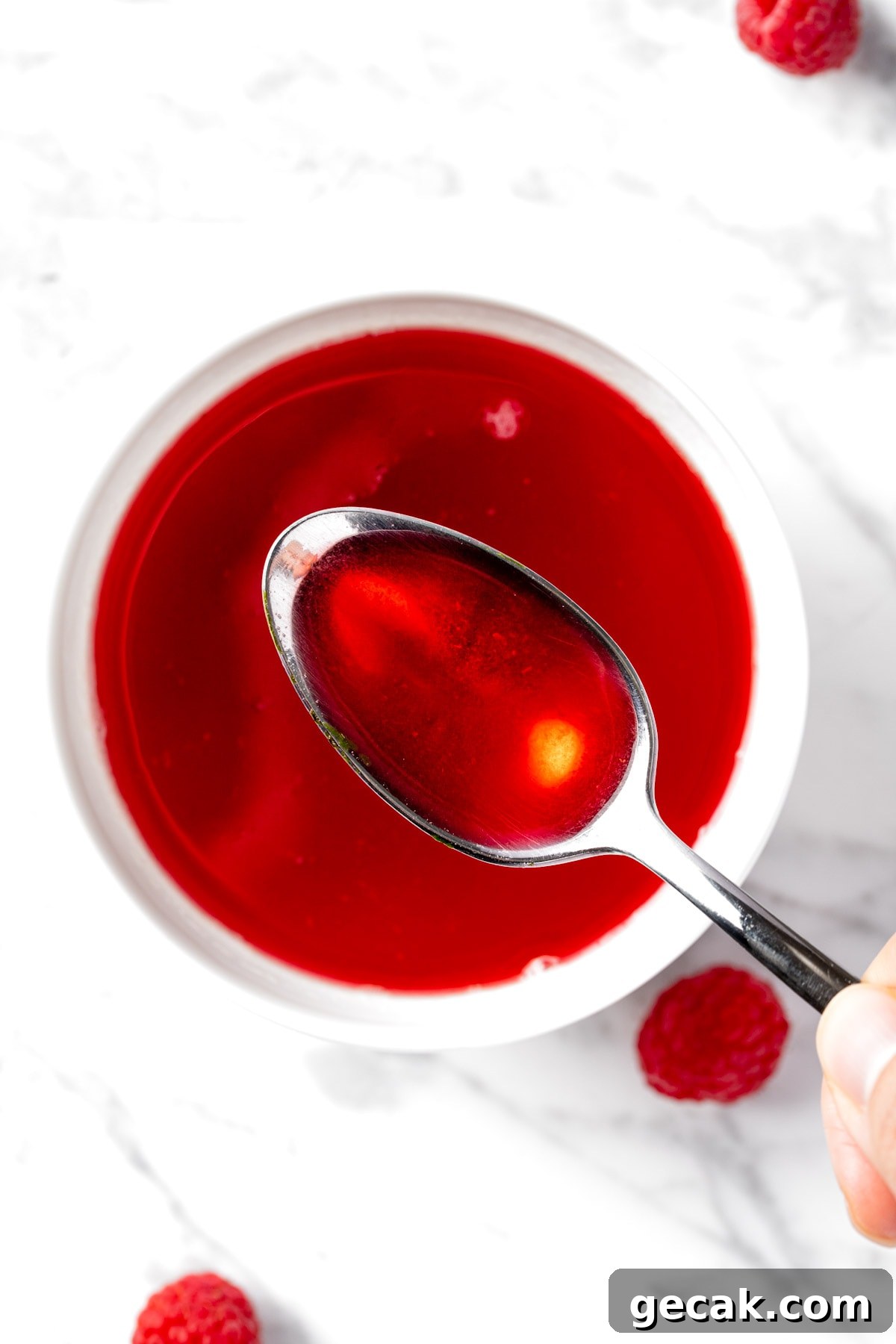 Homemade Raspberry Simple Syrup Recipe for Cocktails & Desserts 2 Overhead view of a spoonful of bright red raspberry syrup, with a white marble background.