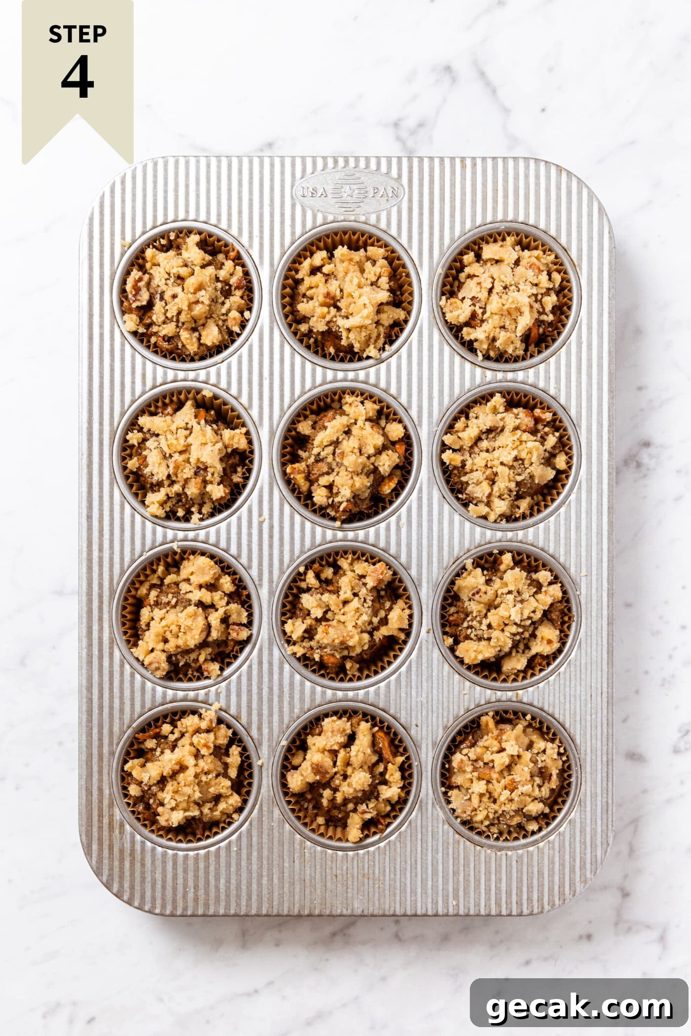 Muffin tin pan resting on white marble surface filled with unbaked carrot muffins with streusel topping.