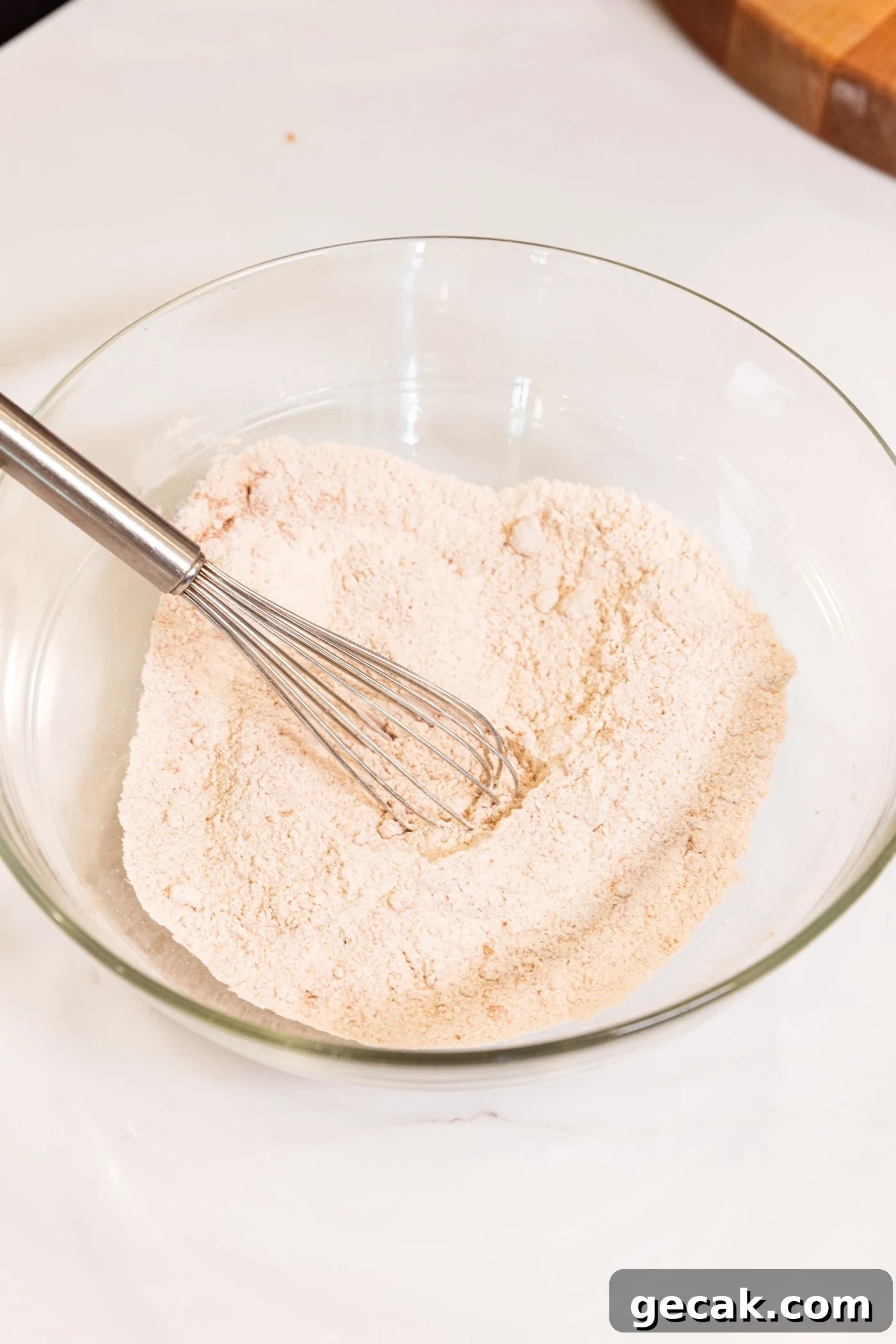 Wire whisk resting against surface of glass bowl with whisked dry ingredients for carrot muffins.