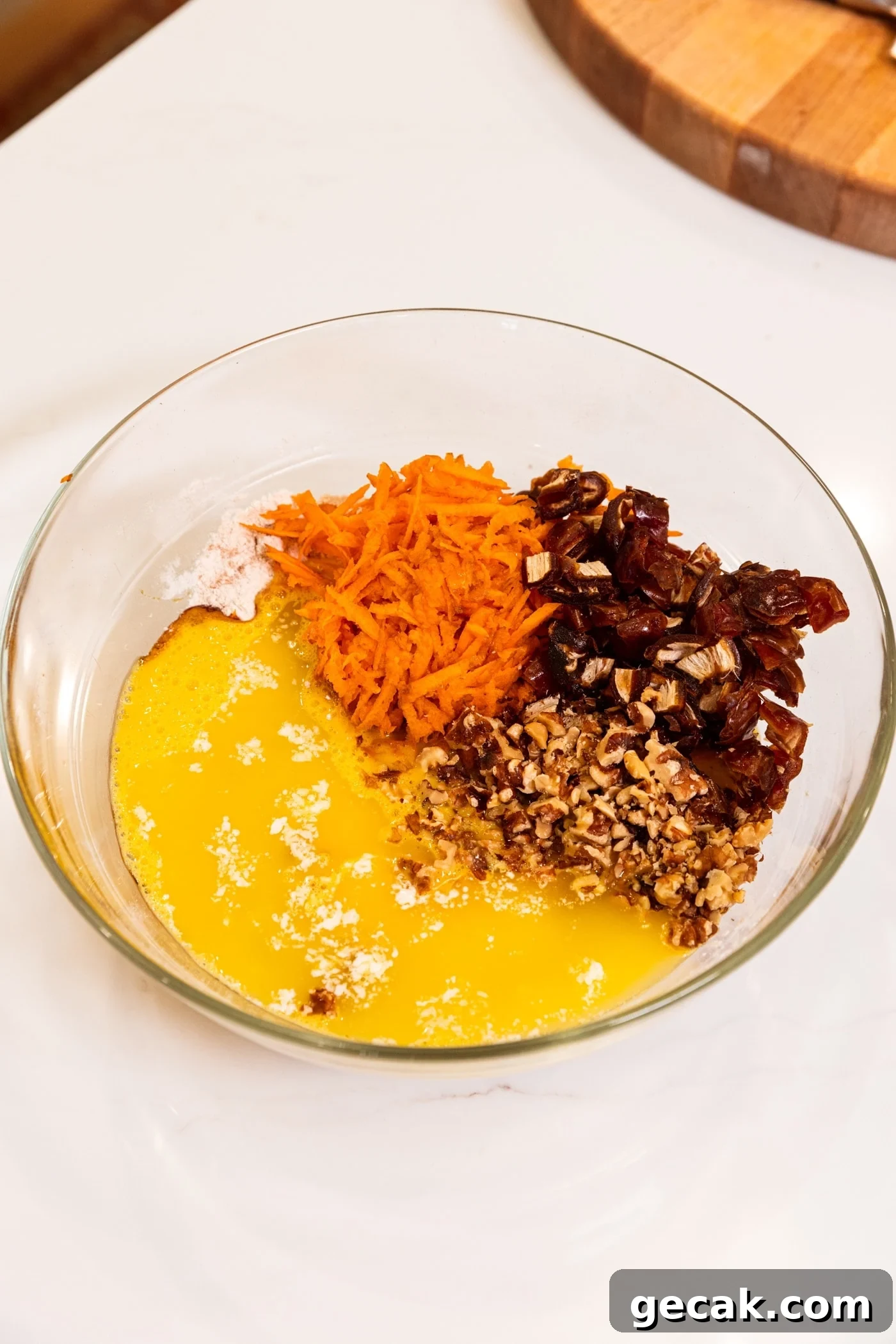 Glass bowl filled with wet ingredients for carrot muffins including shredded carrot, dates, nuts, and eggs.