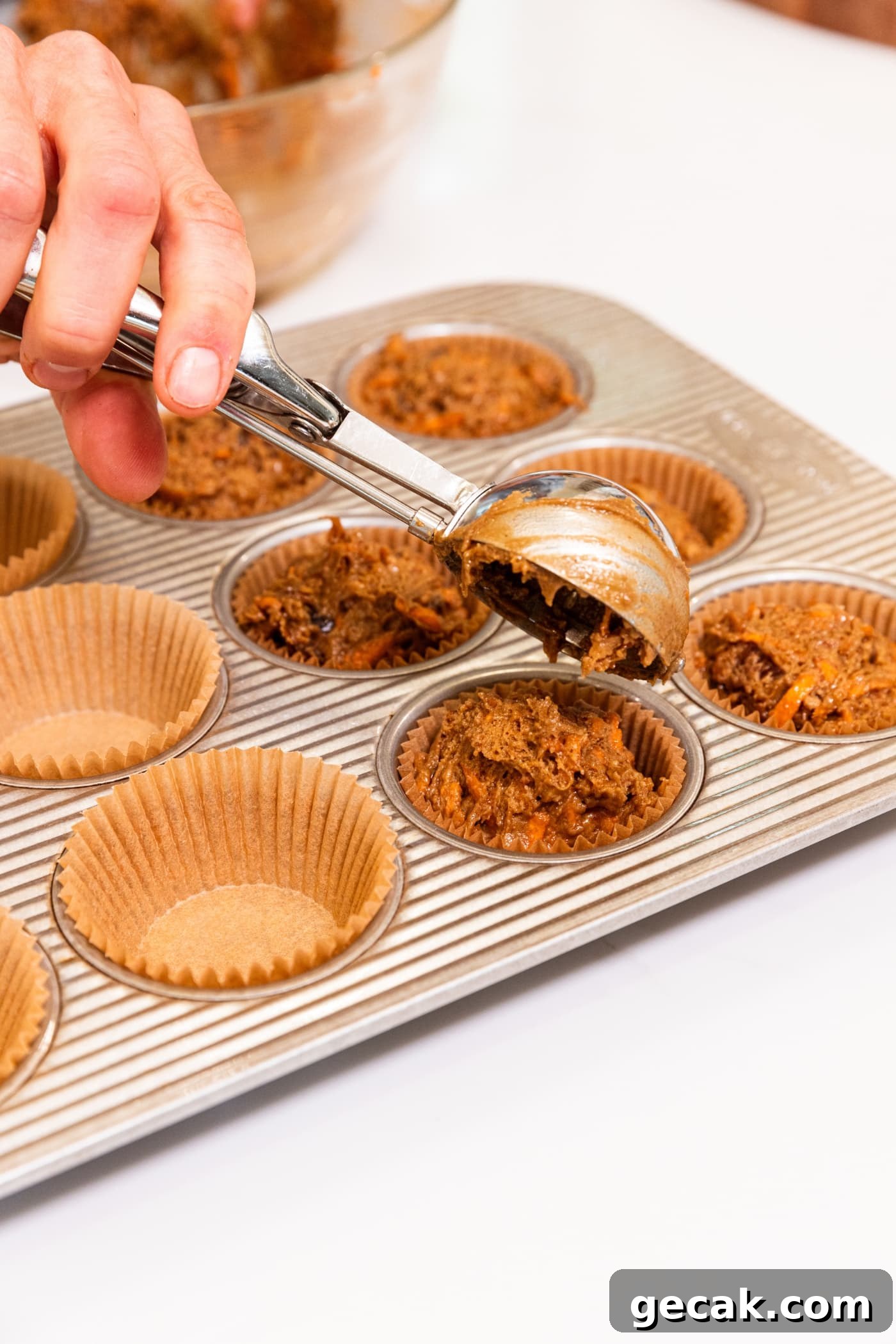 Hand using cookie scoop to place dollops of batter into a muffin pan fitted with muffin cups.