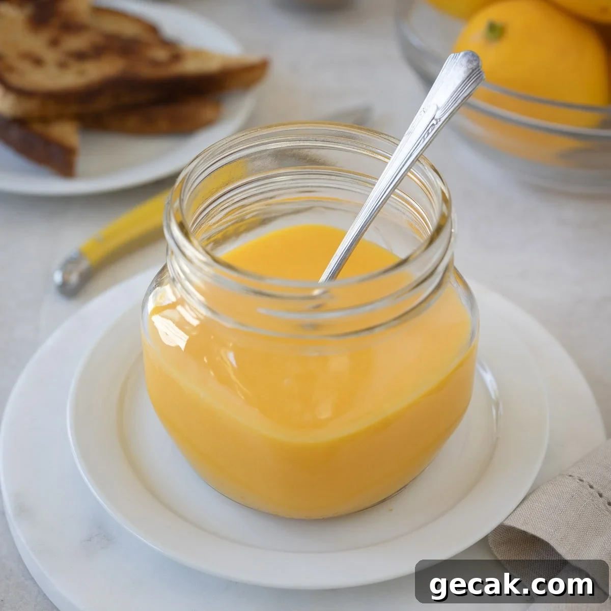 A glass jar of bright yellow lemon curd with a bowl of lemons on the side.