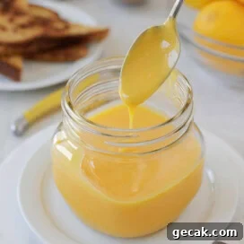 A square jar filled with lemon curd dripping off a silver spoon.