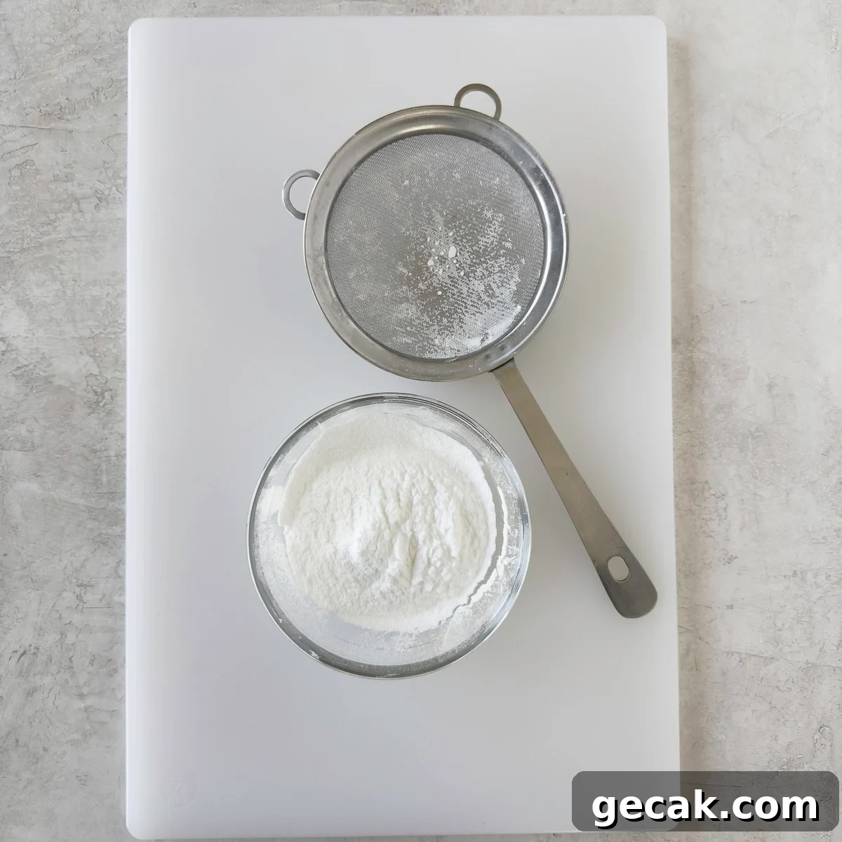 A stainless steel small sieve and sifted powder sugar on a white cutting board.